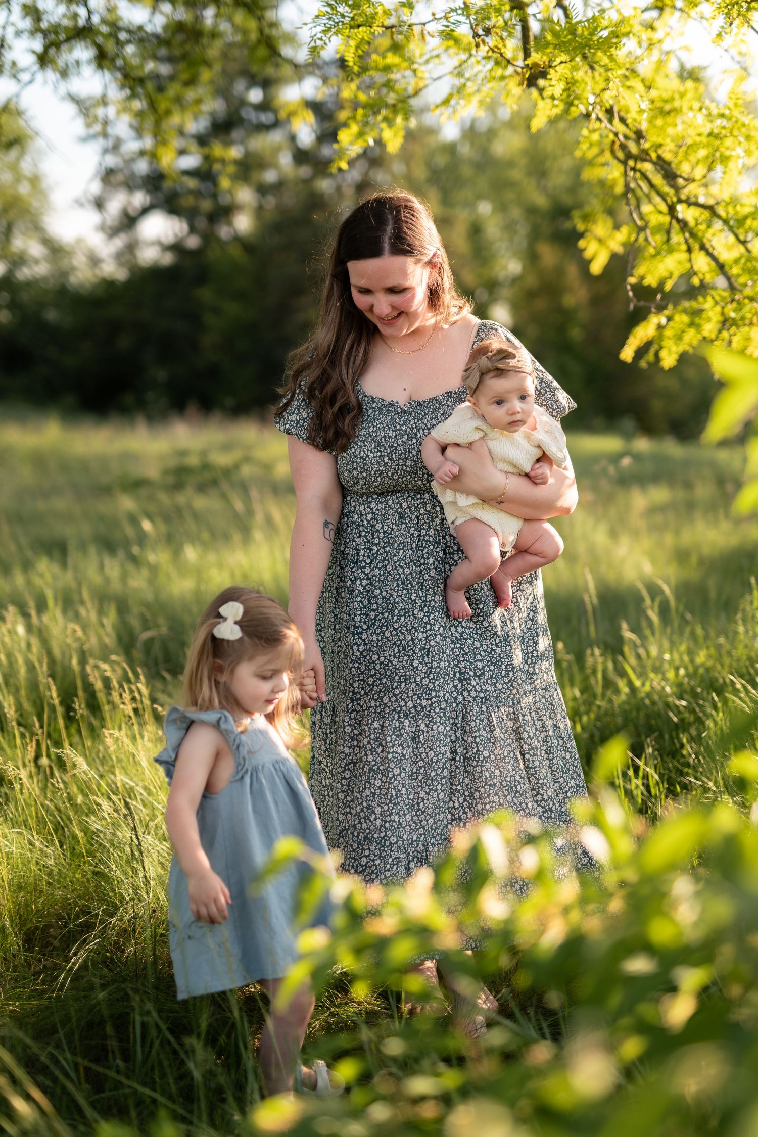 Mother in floral dress holds baby while toddler girl stands beside her in sunny green meadow.