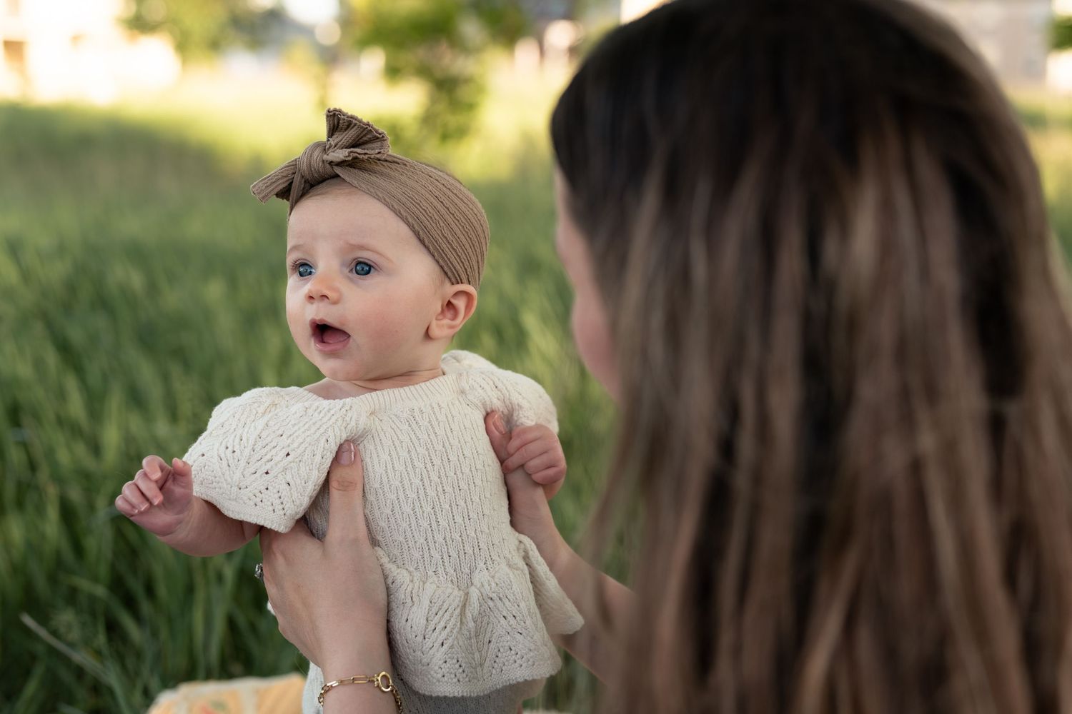 A mother holds her smiling baby girl in a cream knit outfit outdoors in a lush green field.