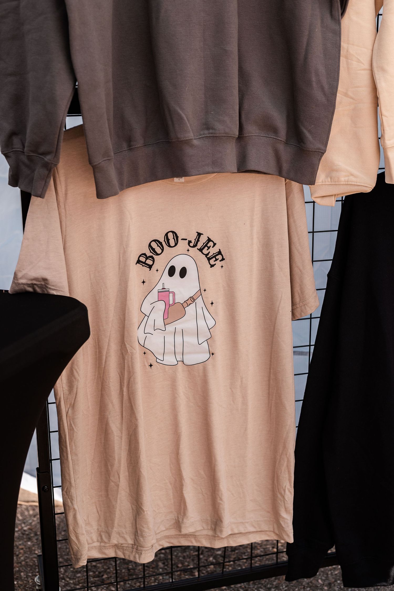 Peach ghost graphic tee reading Boo-Jee hanging at an outdoor market beside brown sweatshirt.