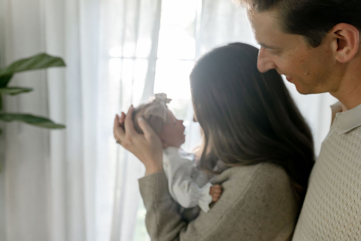 A couple shares a tender moment holding their newborn baby near a sunlit window at home.