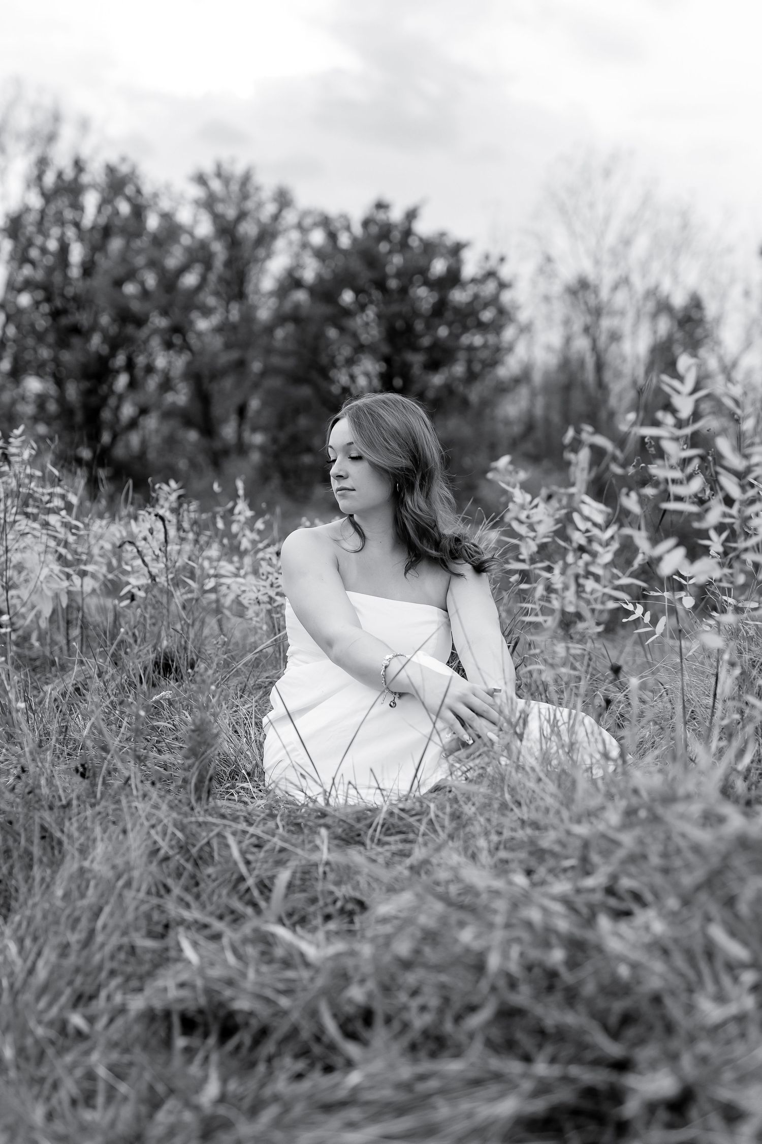A woman in a white dress sits gracefully in tall grass, captured in a serene black and white photo.