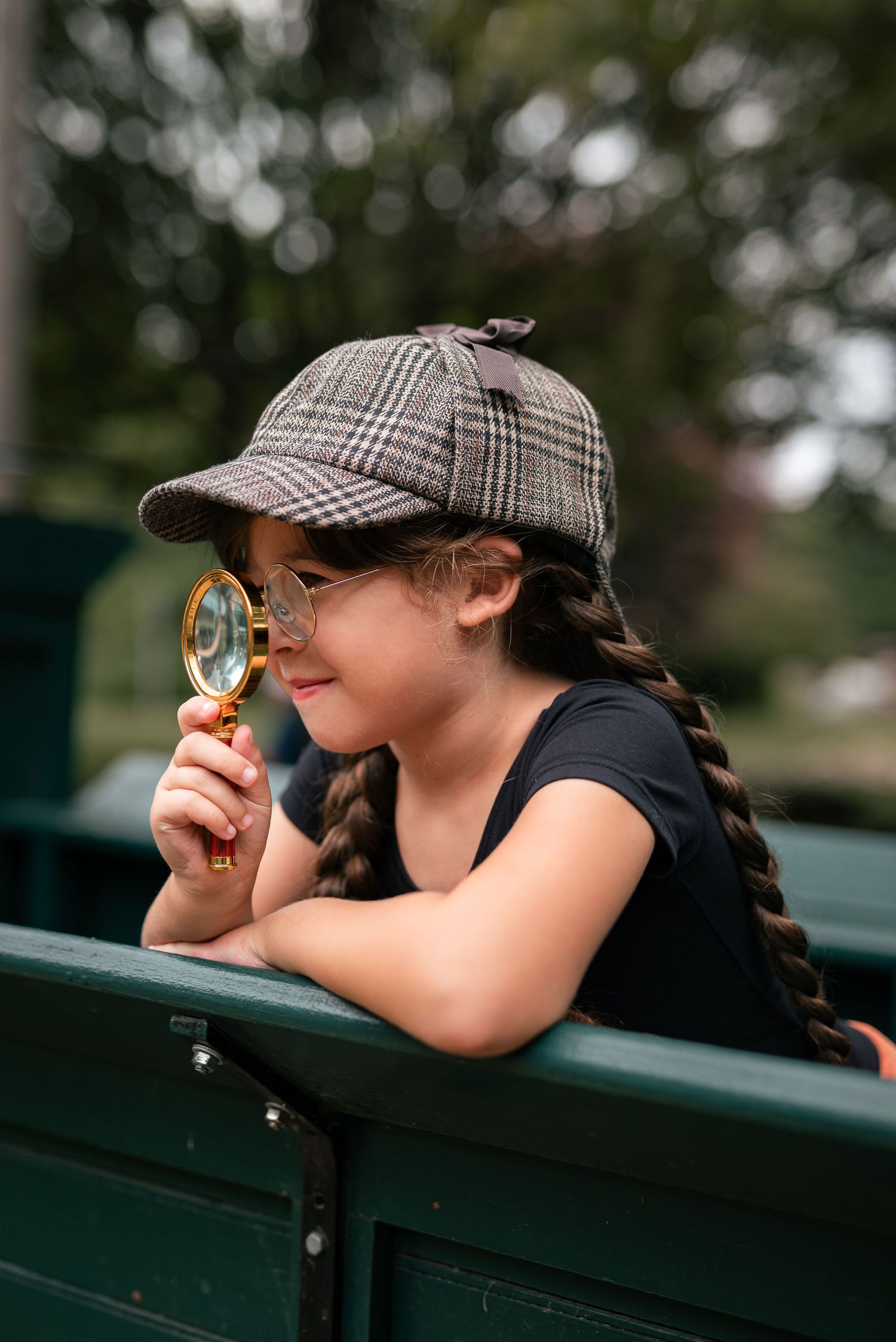 Young girl wearing plaid detective cap holds magnifying glass outdoors, playing inspector near green bench.