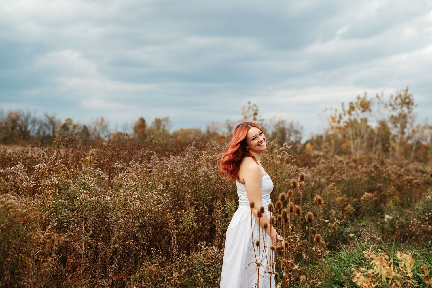Red-haired woman in white dress standing in autumn field under cloudy sky.