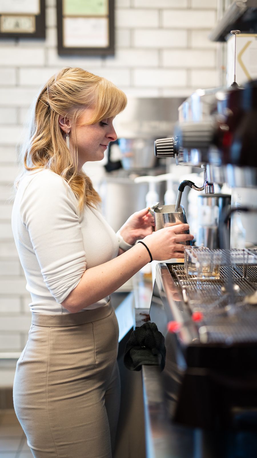 Barista in white shirt and tan apron prepares coffee drinks at a cafe counter.