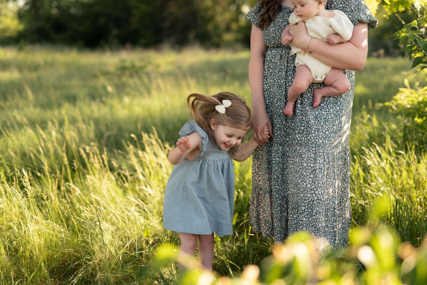 Mother in floral dress holds baby while toddler girl in blue dress plays in sunny green meadow.
