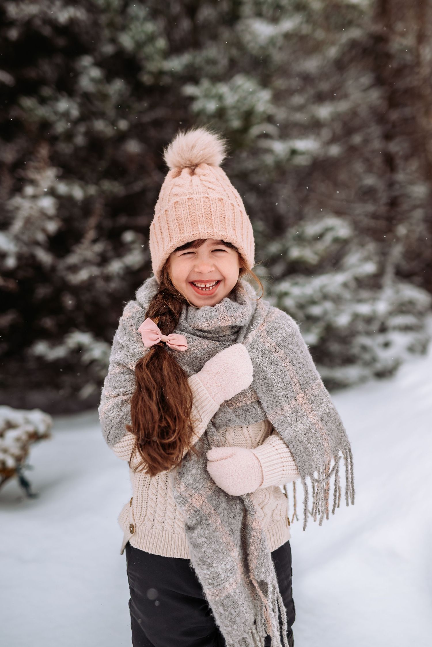 A person wearing a pink knit hat, gray scarf and mittens smiles while enjoying a snowy winter day outdoors.