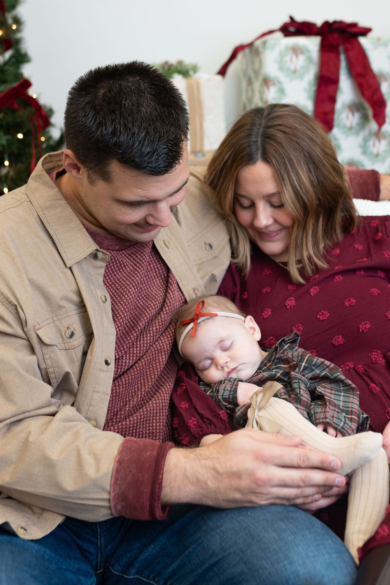 A couple smiles at their sleeping newborn baby during a Christmas photo session with gifts and tree in background.