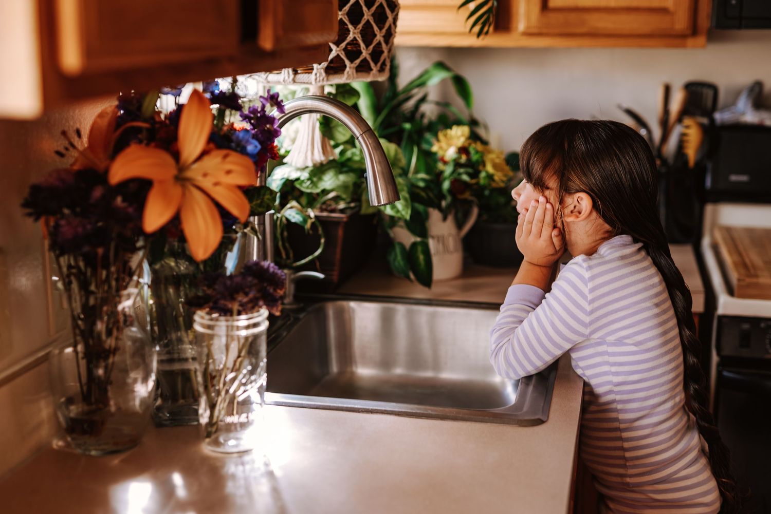 Person sitting at kitchen counter with hands covering face next to vase of orange lilies in warm indoor lighting.