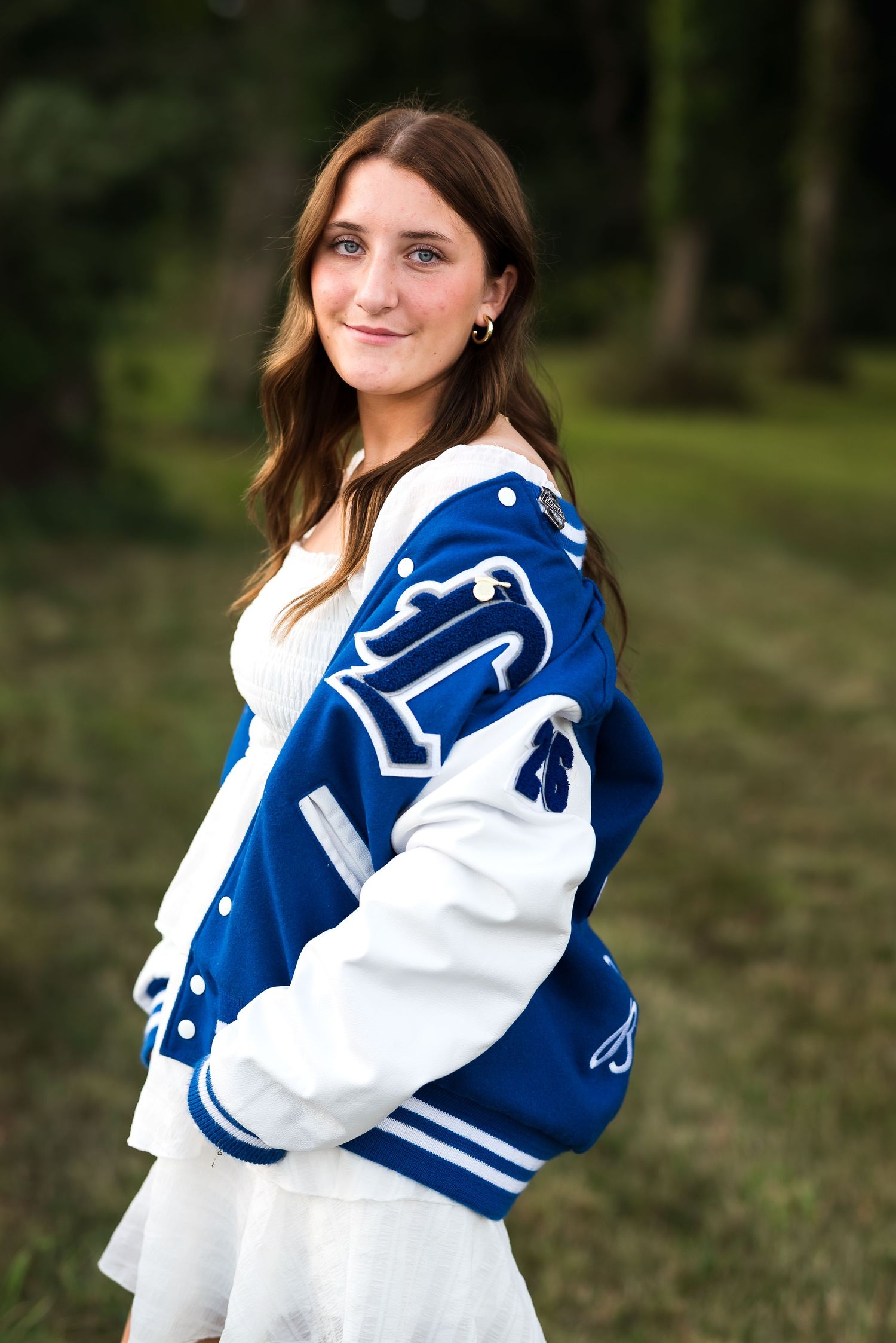A person wearing a blue and white varsity jacket poses outdoors in a grassy area with trees in the background.