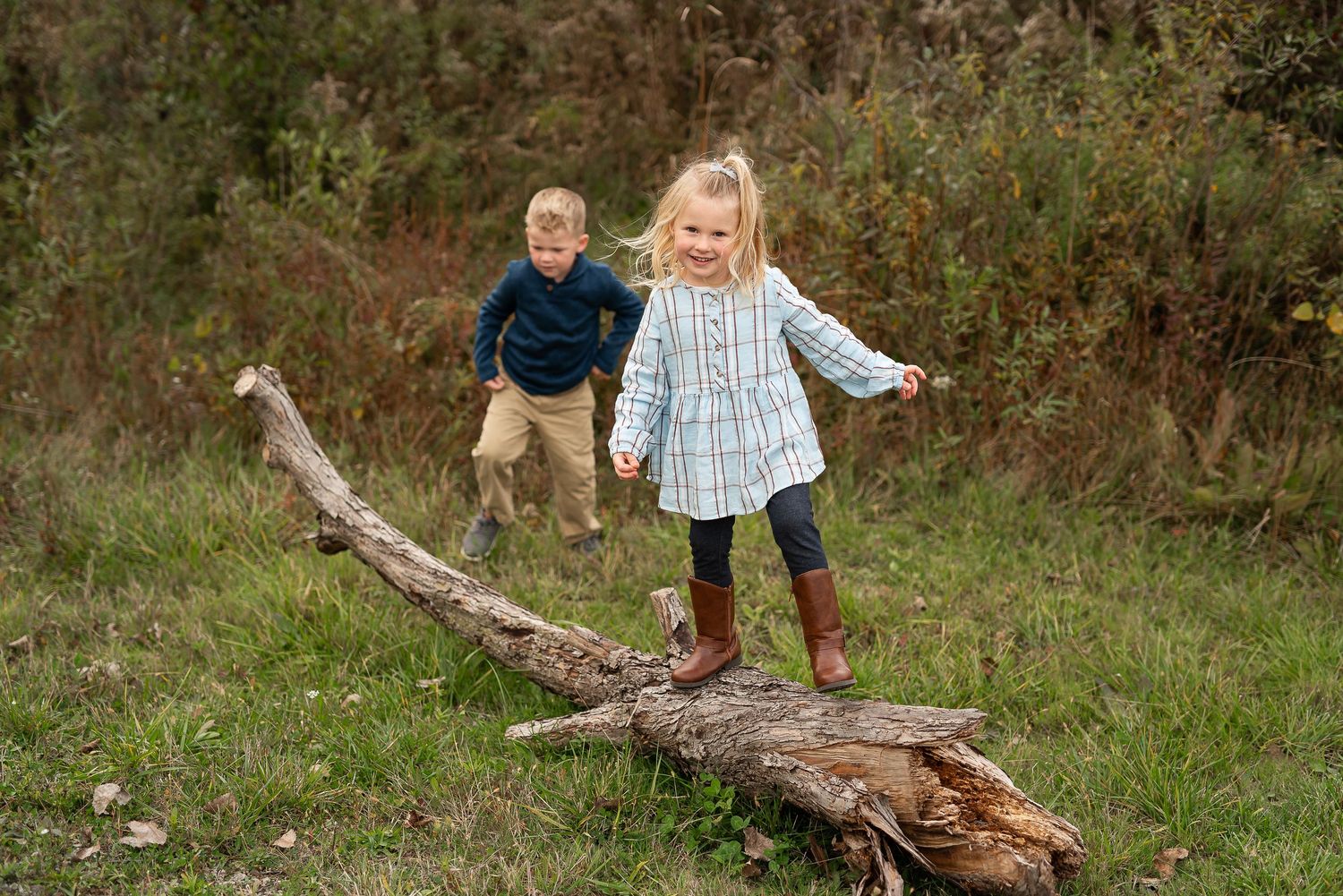 Two young children playing outdoors, balancing on a fallen log in a green meadow.