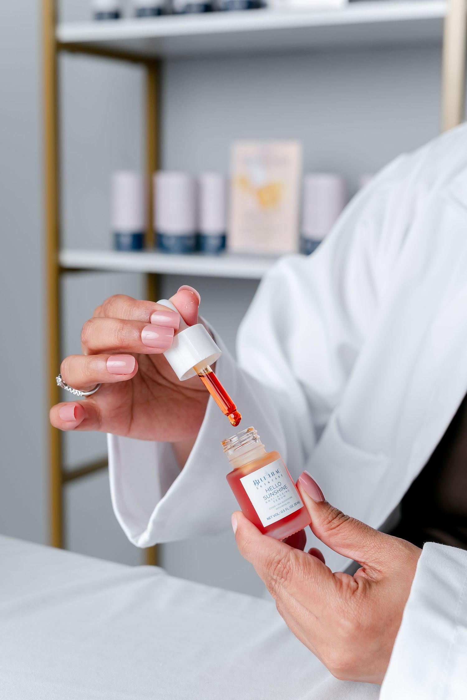 Esthetician in white coat holding a dropper bottle of facial serum in a professional skincare studio.