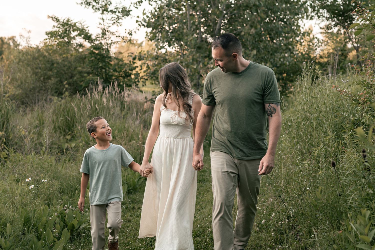 Family of three walking through lush green meadow, parents holding child's hands at golden hour.