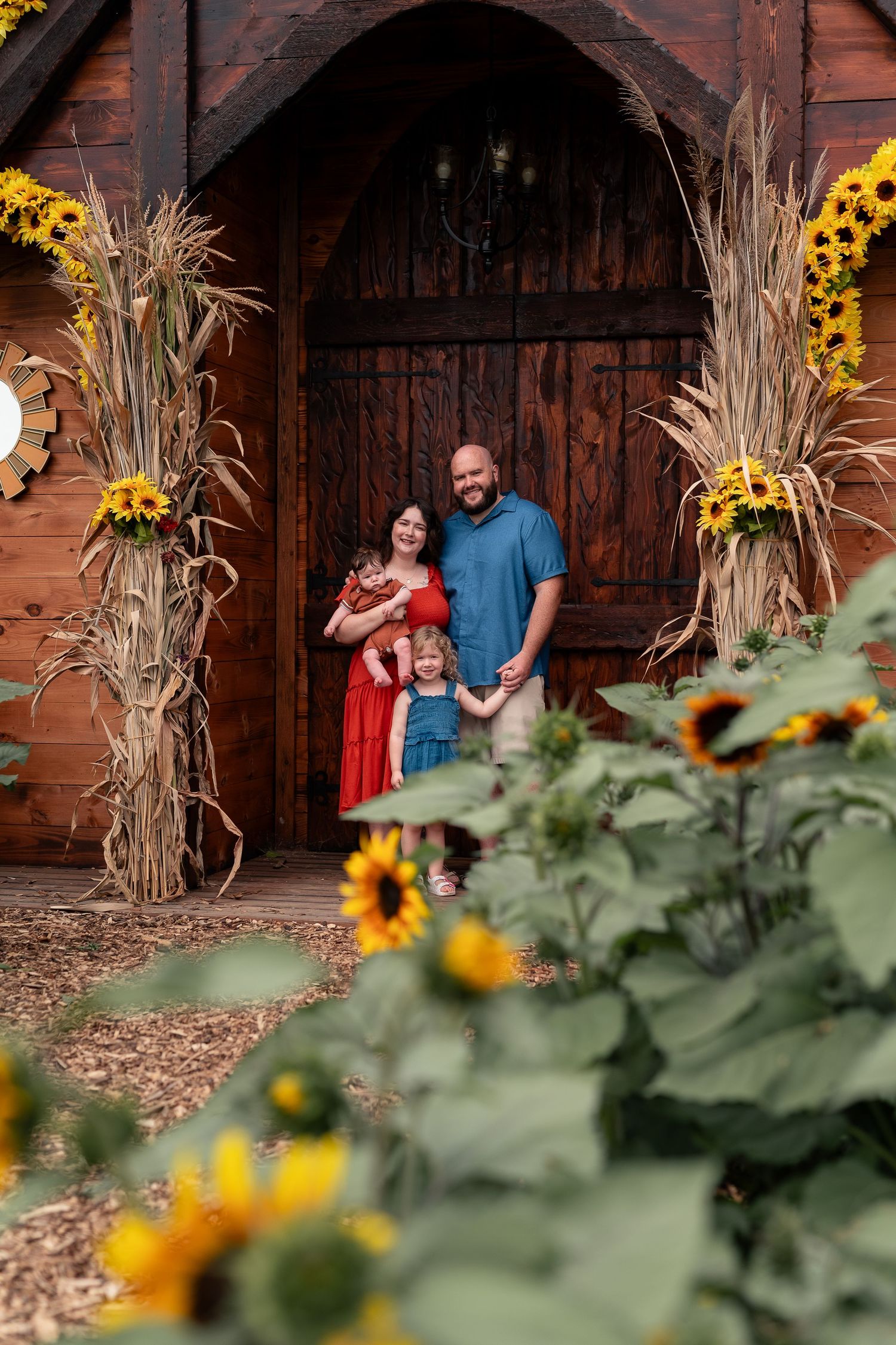 Family of four posing in front of rustic wooden barn doors surrounded by sunflowers and corn stalks decorations.