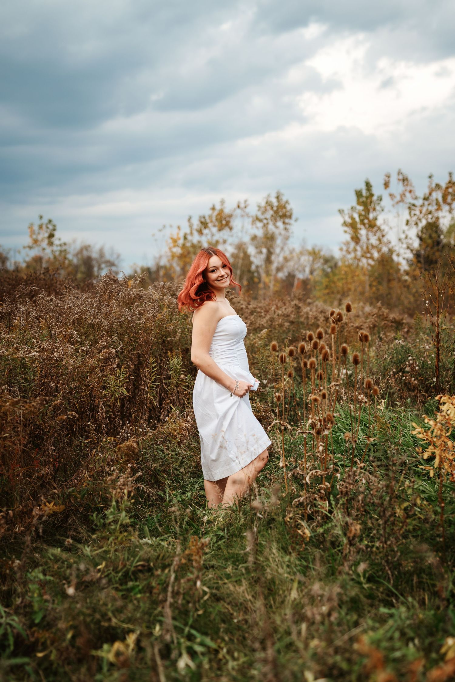 Young woman with red hair in white dress standing in autumn field with dramatic cloudy sky.