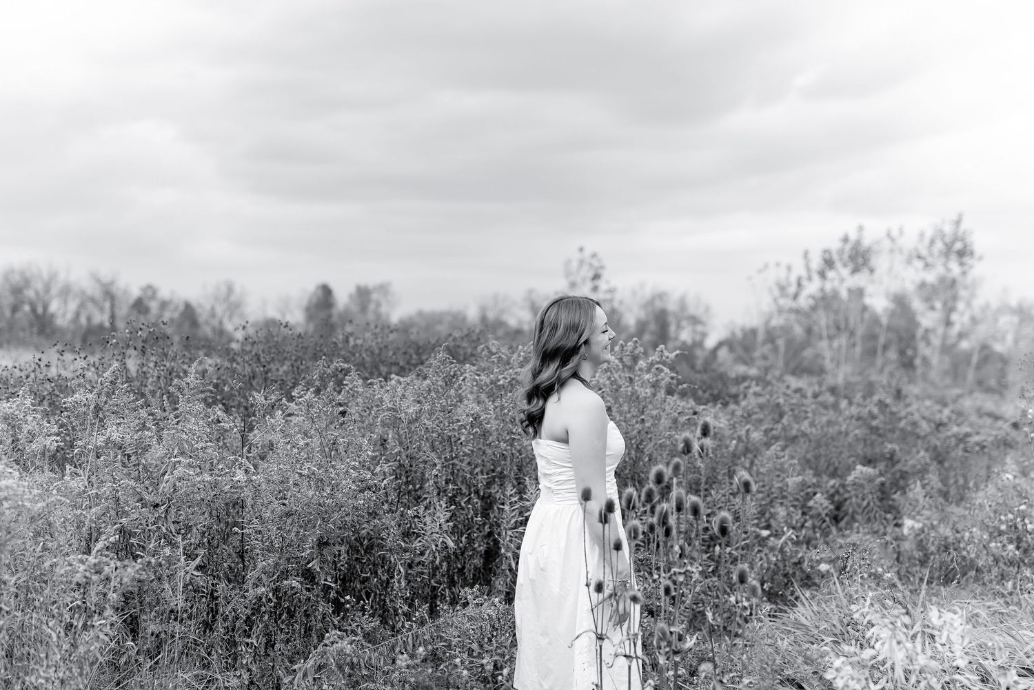 Black and white photo of a woman in a white dress standing in a field of wildflowers under a cloudy sky.