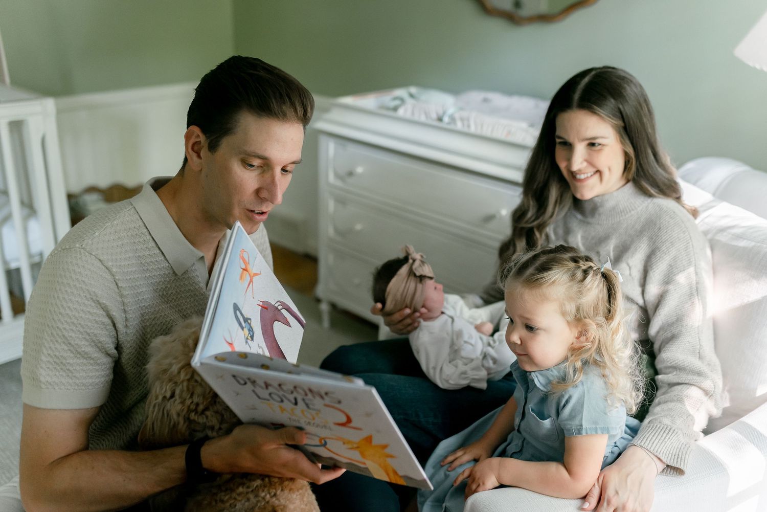 Family reads a colorful picture book together in a cozy nursery bedroom setting.