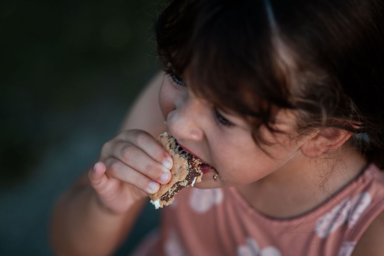 Young girl with dark hair eating a granola bar snack, close-up candid photo of child enjoying food outdoors.
