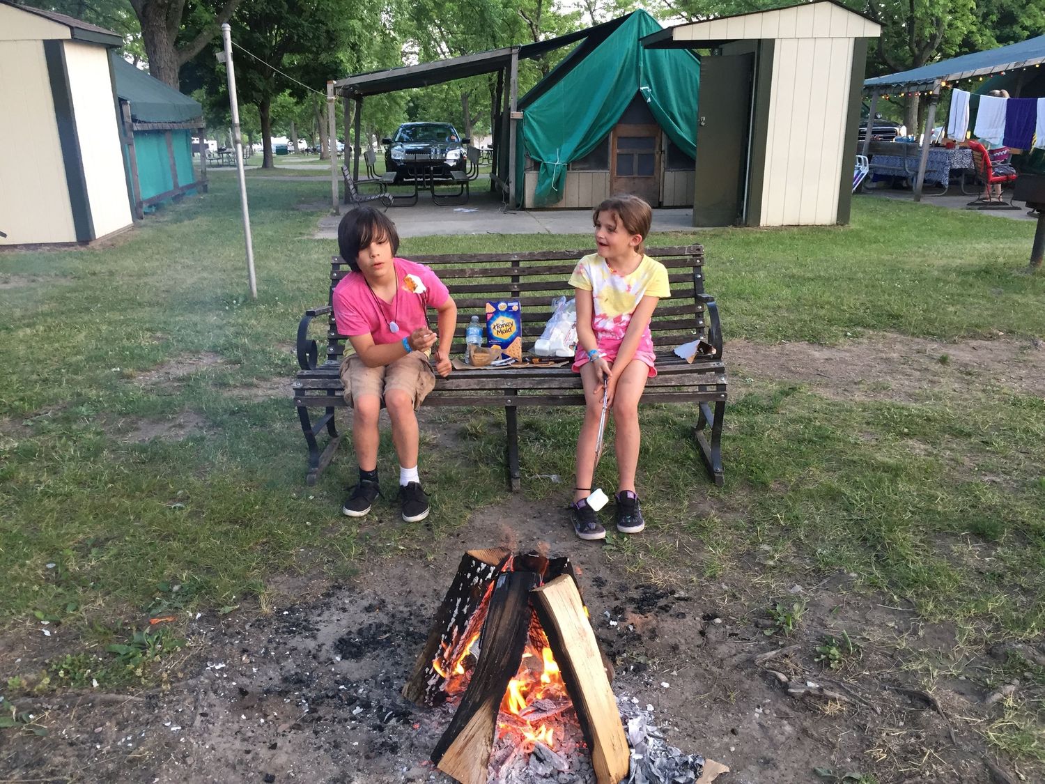 Two children sit on a bench roasting marshmallows over a campfire at a campground with tents in the background.