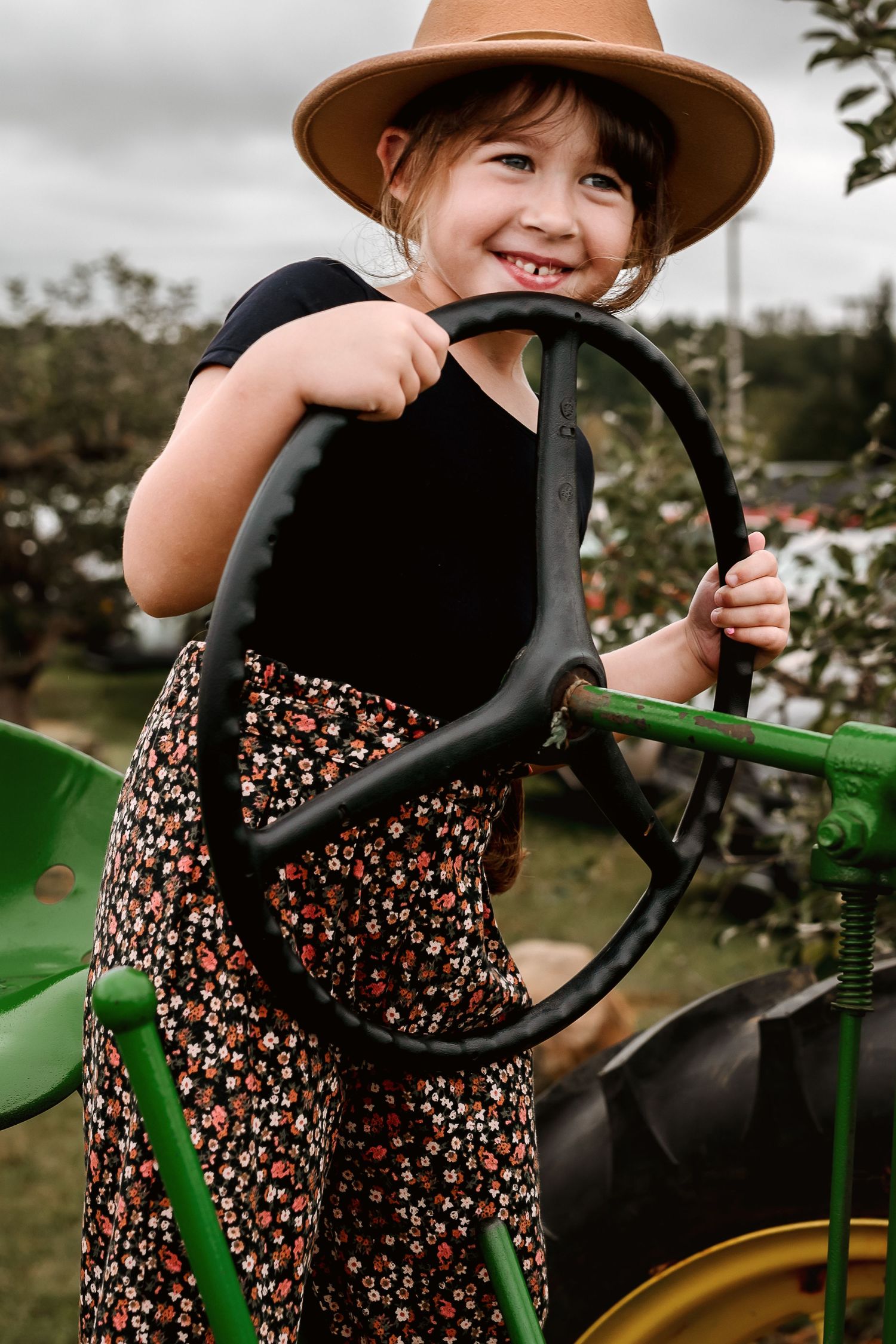 A child in a brown hat and floral dress smiles while holding onto the steering wheel of a vintage green tractor.