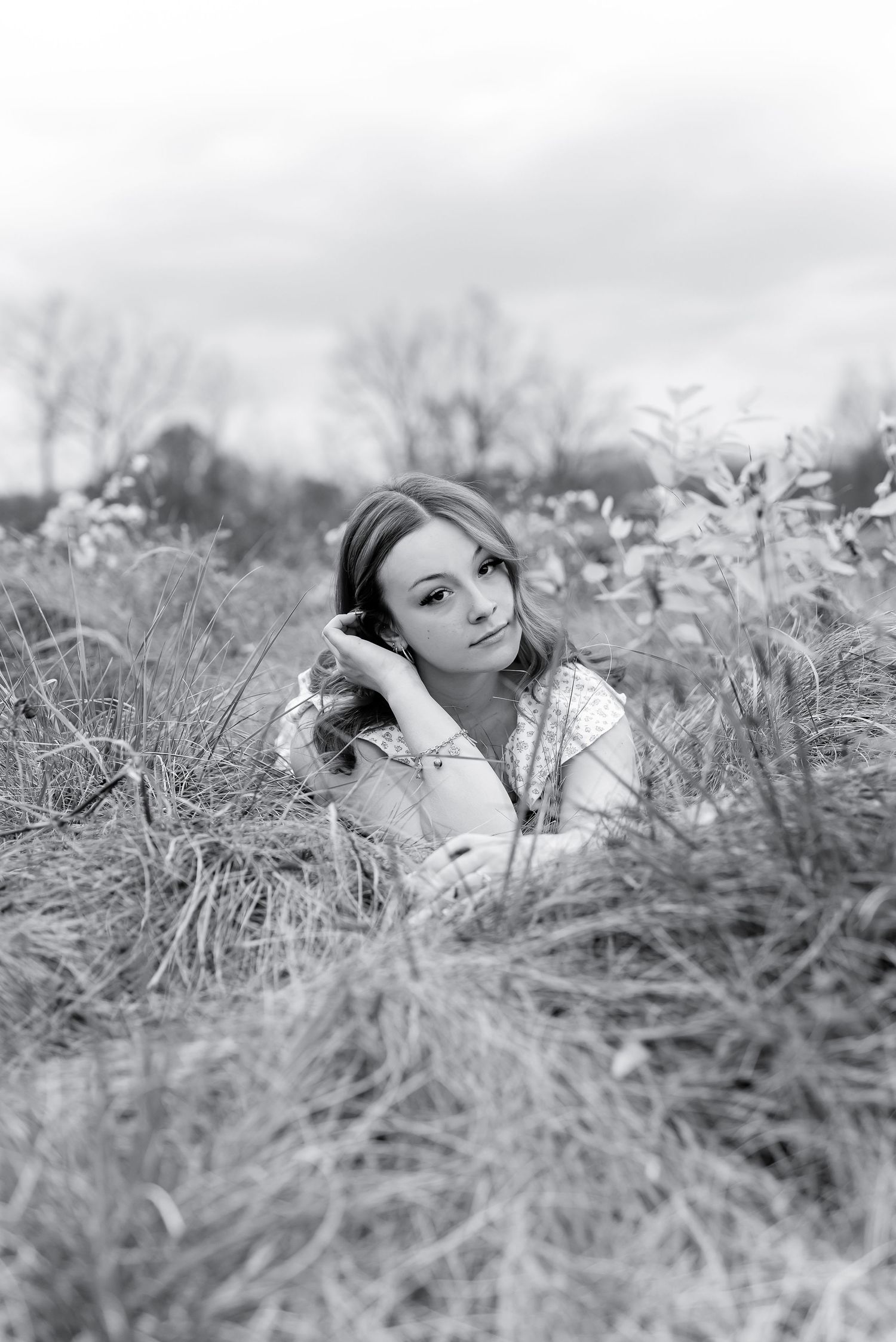 Black and white portrait of young woman lying in tall grass field looking at camera.