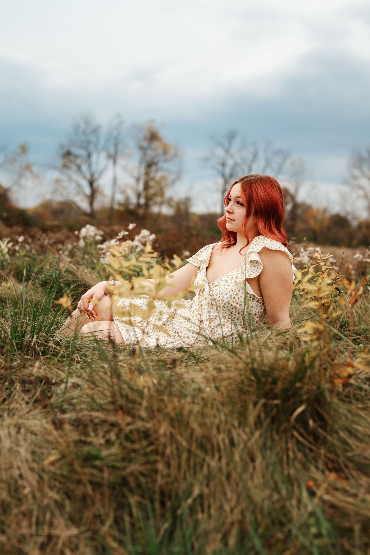 Red-haired woman in floral dress sitting in tall grass field with wildflowers and trees under cloudy sky.