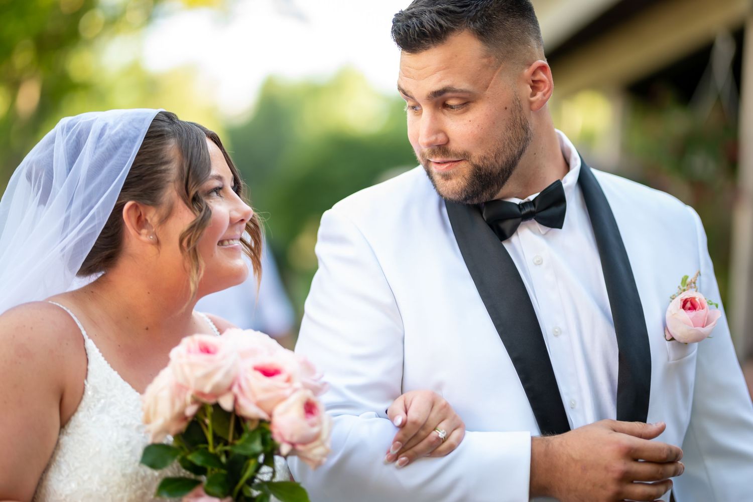 A joyful couple celebrating on their wedding day, wearing elegant white attire with a bouquet of pink roses.