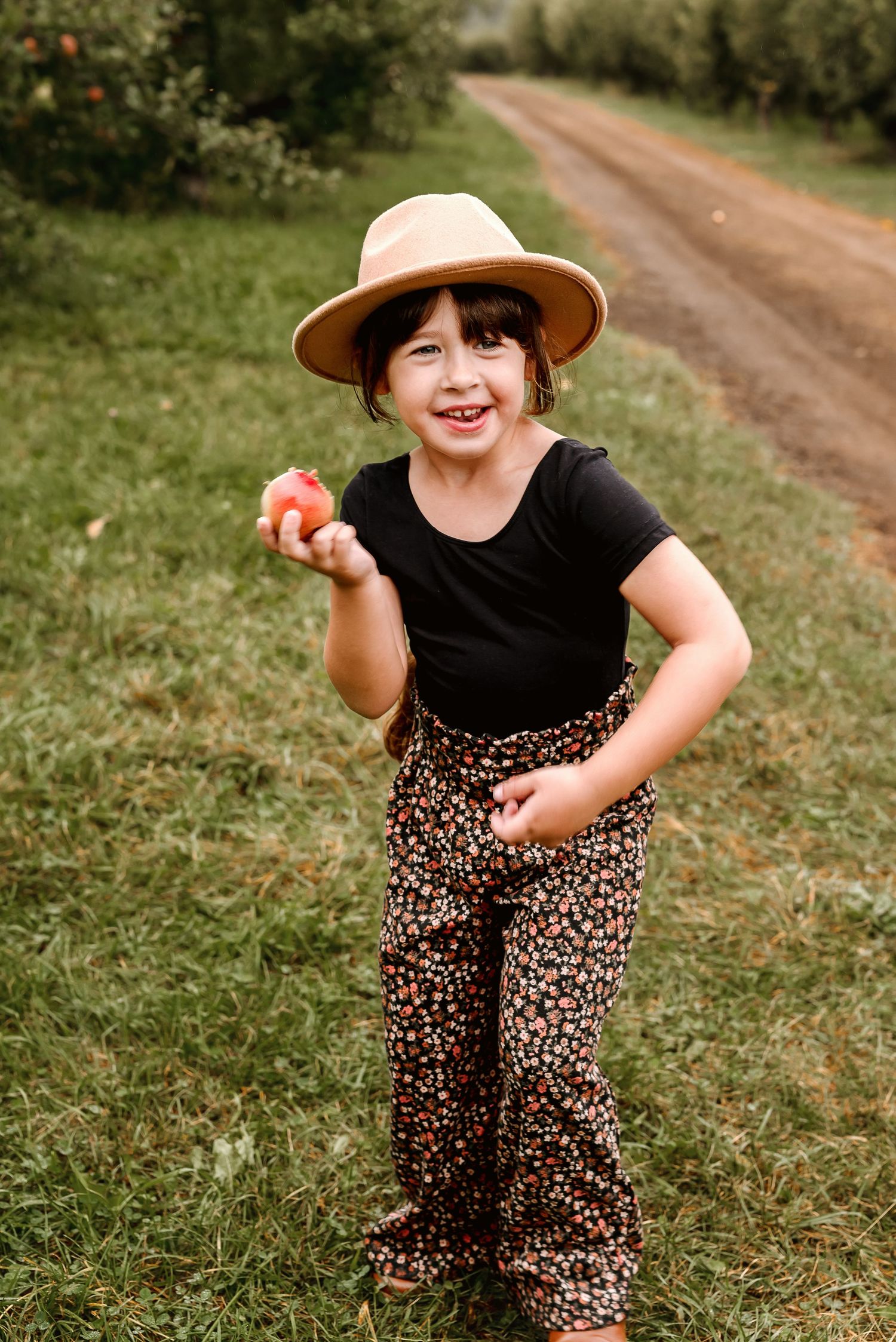 A child in a black shirt and leopard print pants plays outdoors on a dirt path surrounded by green grass.