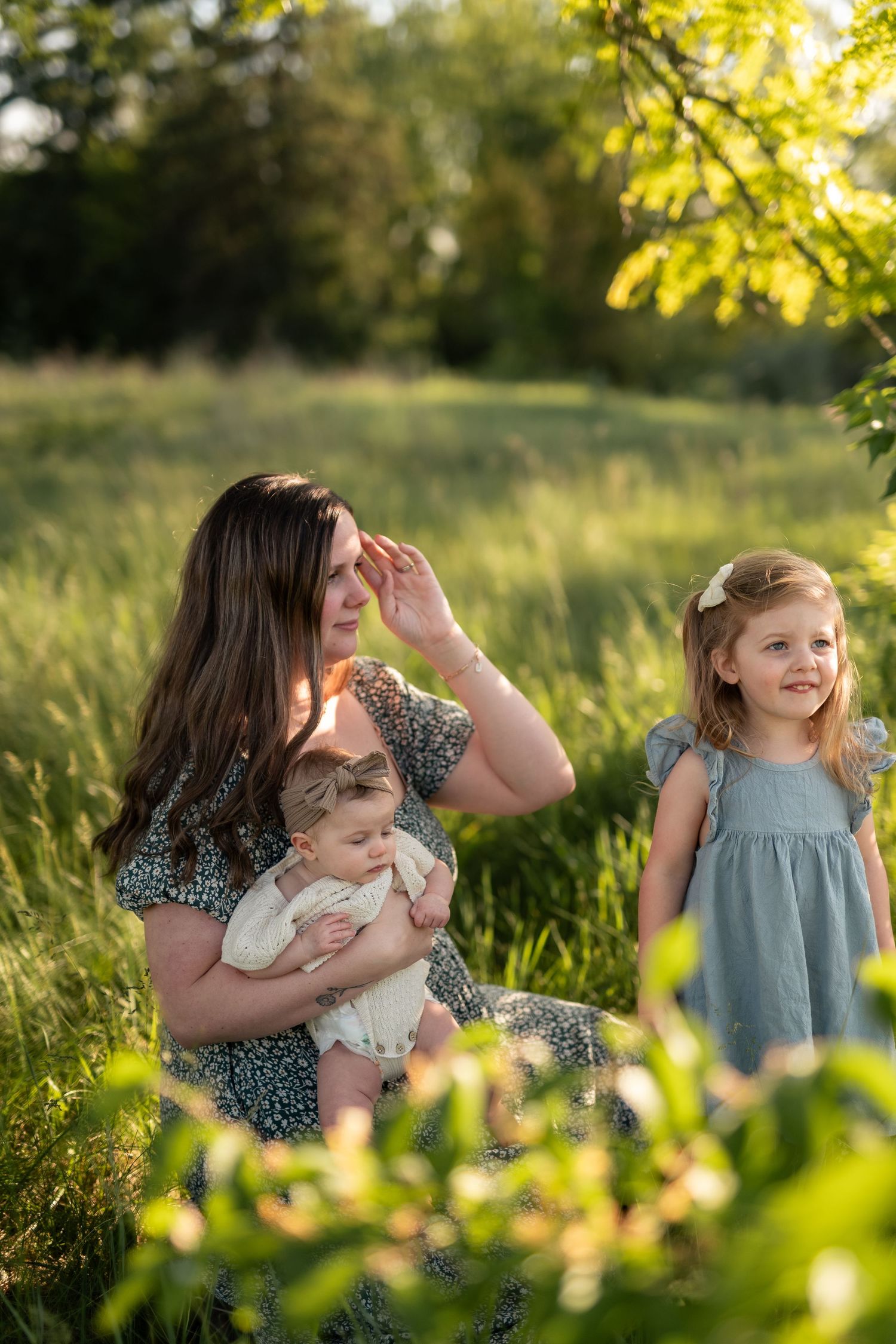 A mother sits in a sunny green field holding her baby while her young daughter in a blue dress stands smiling beside her.
