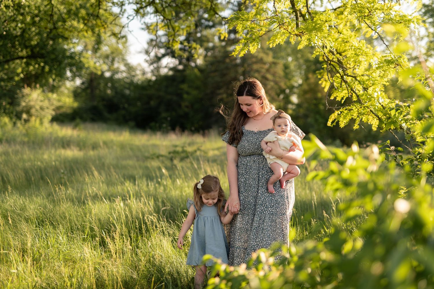 Mother holding baby walks with young daughter through lush green meadow in golden sunlight.