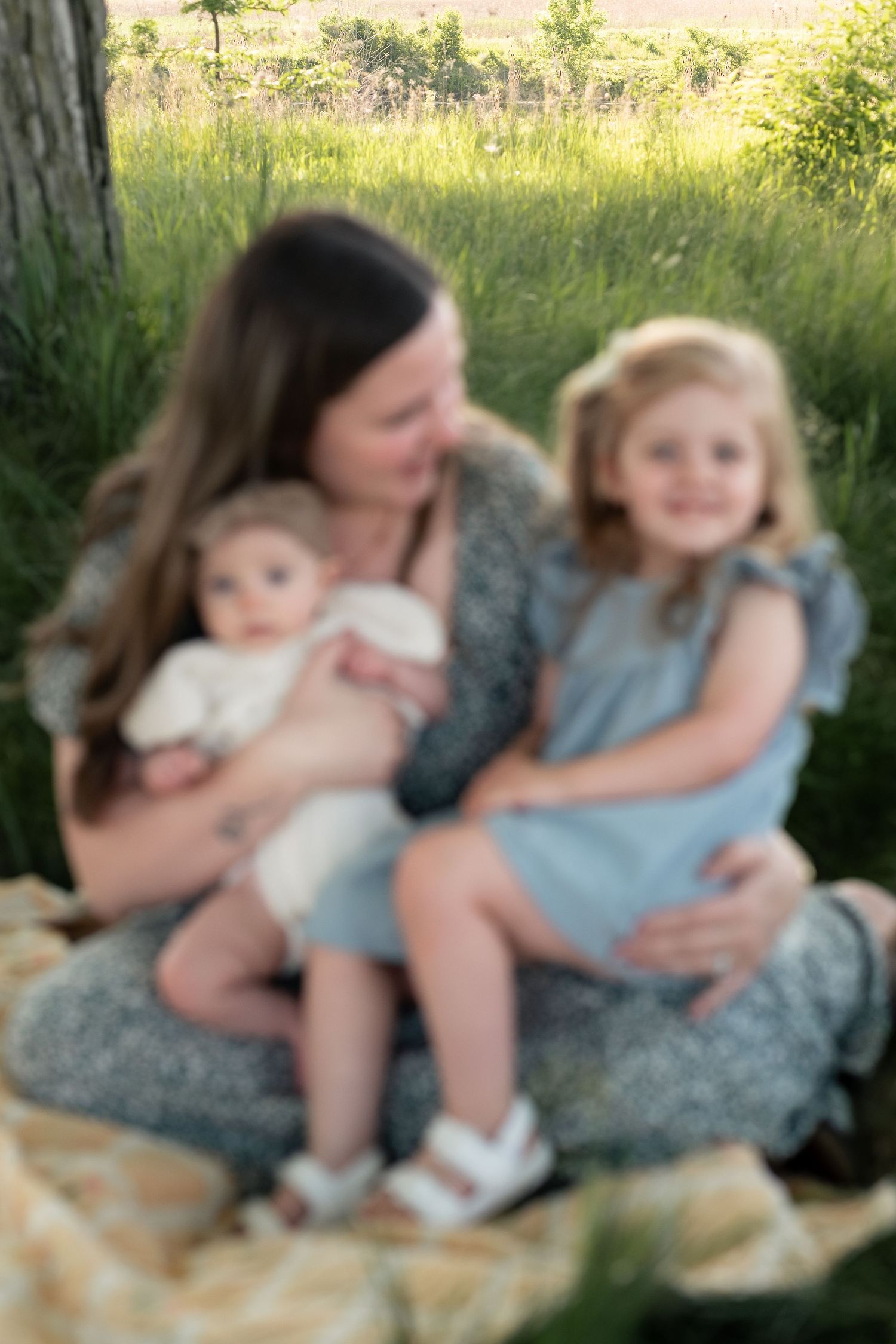 A mother sits on a blanket in a green field, lovingly holding her baby and young blonde daughter in blue dress.