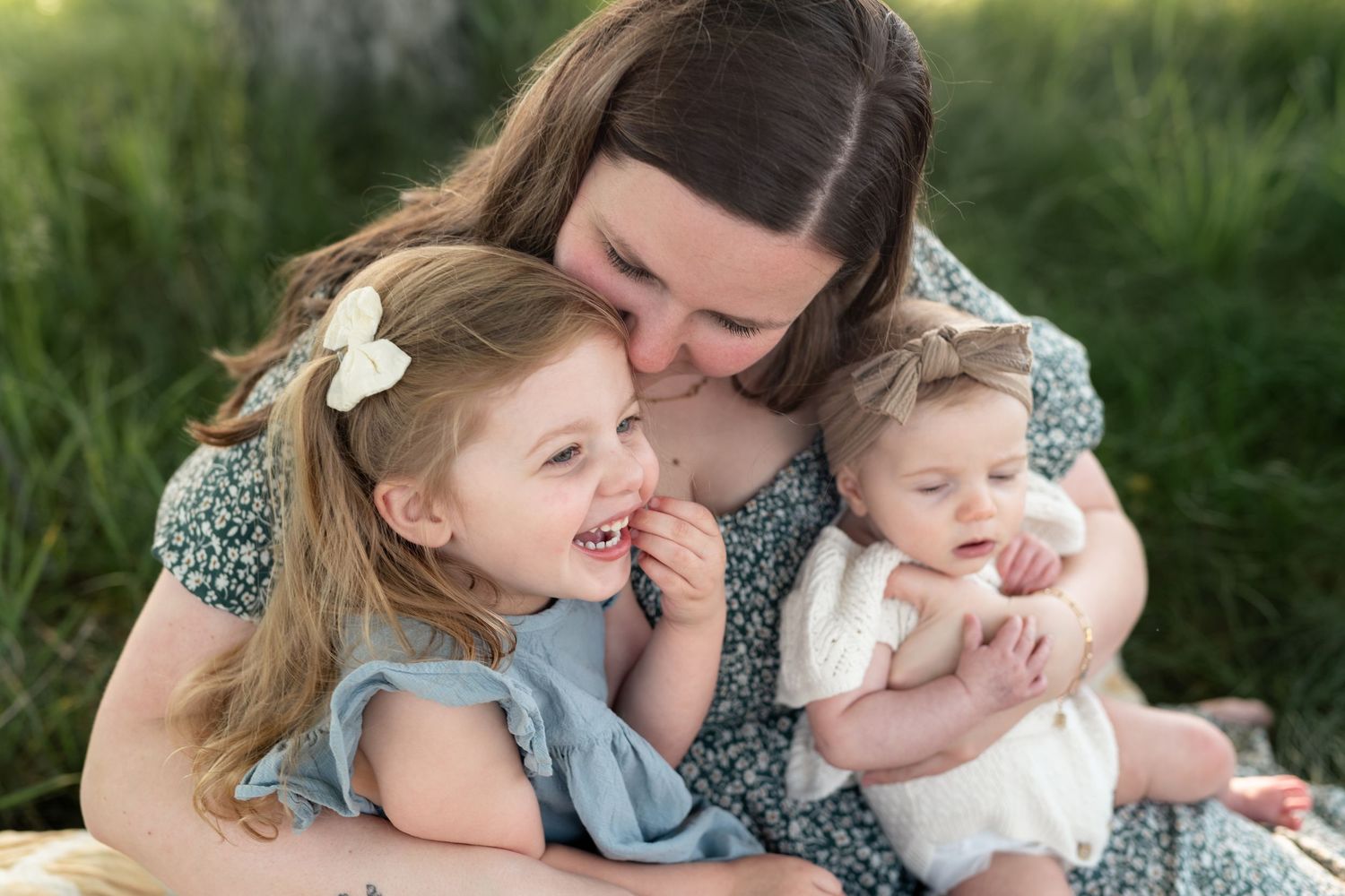 A mother tenderly kisses her smiling toddler girl while holding her baby outdoors in a green field.