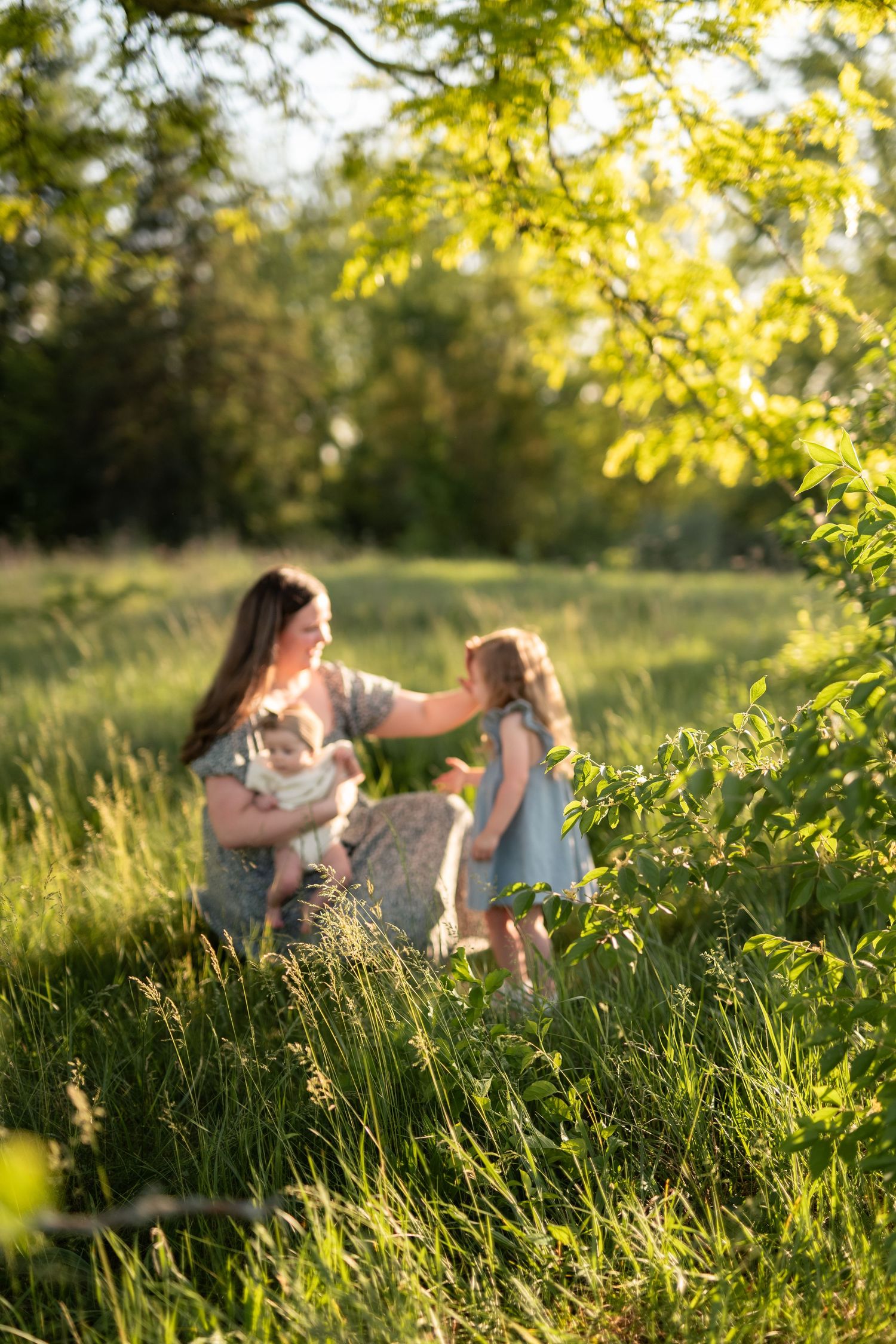Mother holding baby while young daughter reaches out to touch sibling in sunny green meadow.