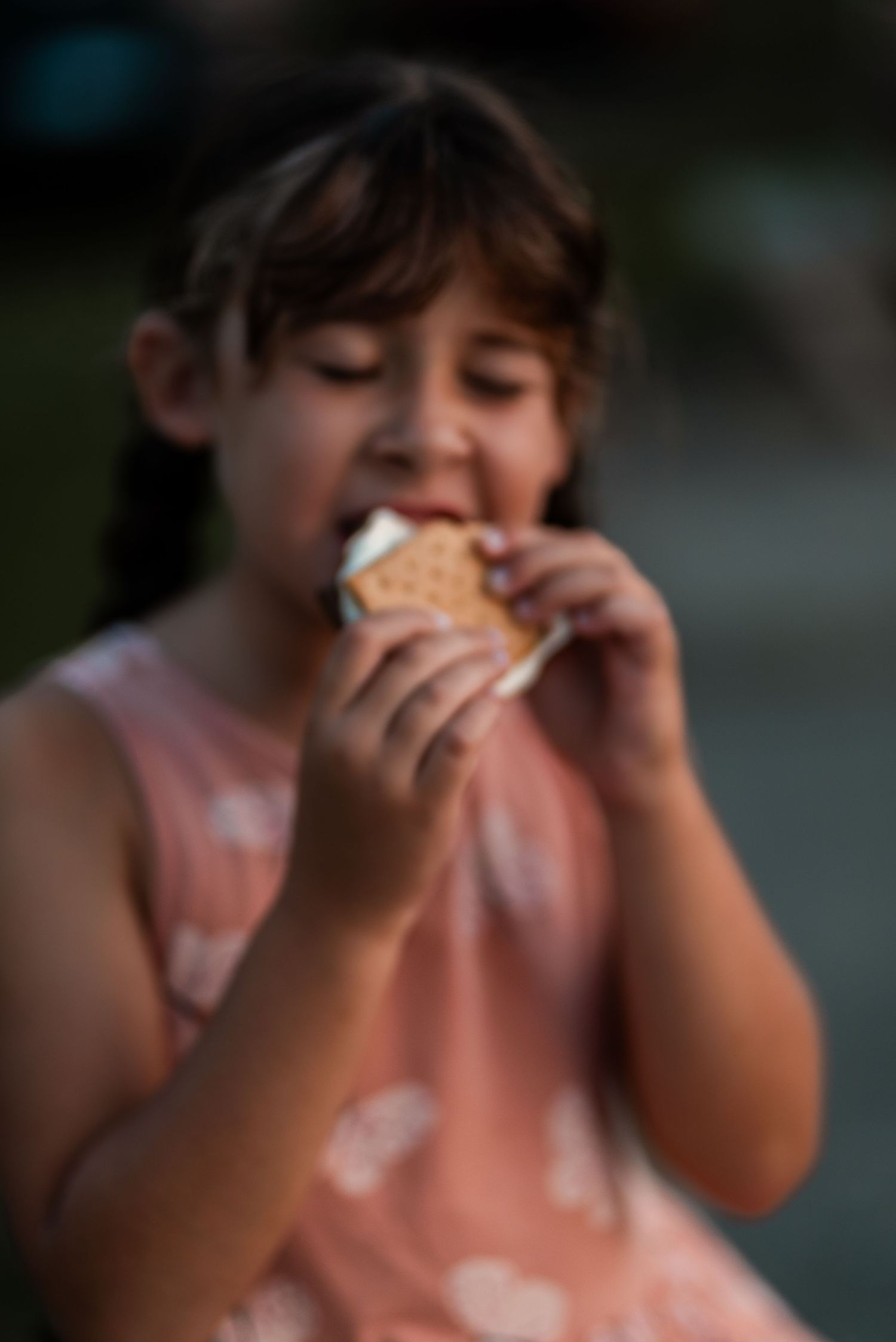 A young girl in a pink dress enjoys eating a s'more outdoors, with eyes closed in delight.