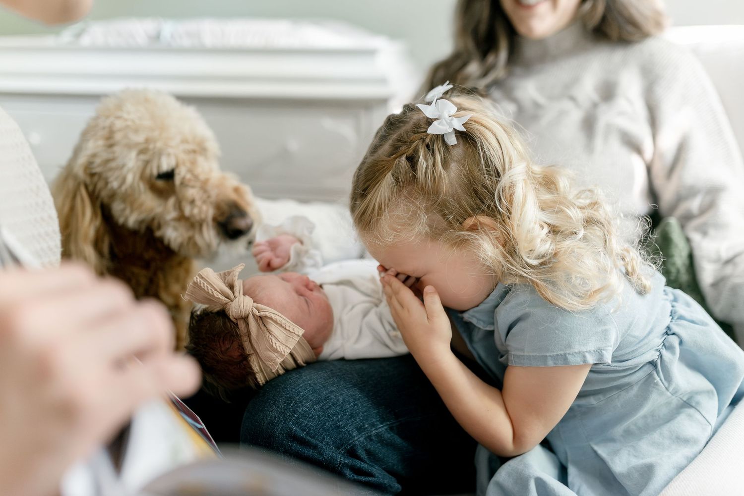 A toddler girl kisses a swaddled newborn baby while a golden doodle dog watches on a cozy sofa.