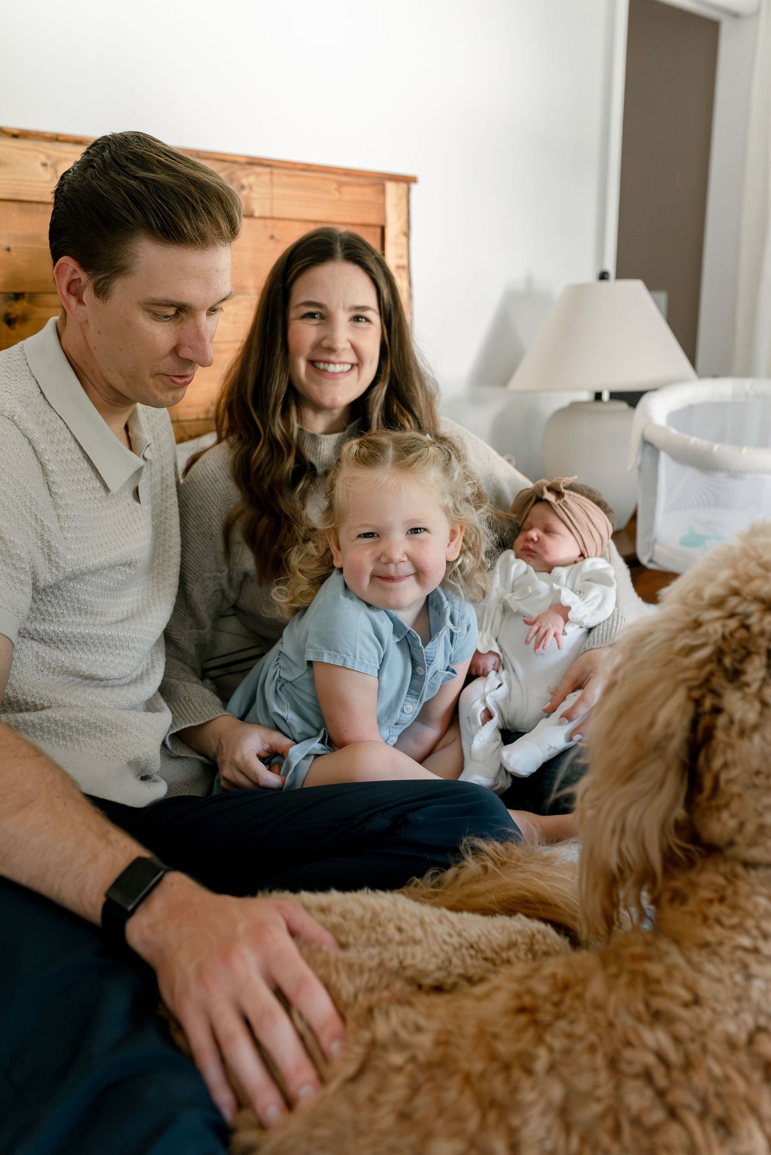 A happy family with two parents, a toddler girl, and a newborn baby sit on a bed with a golden retriever dog nearby.
