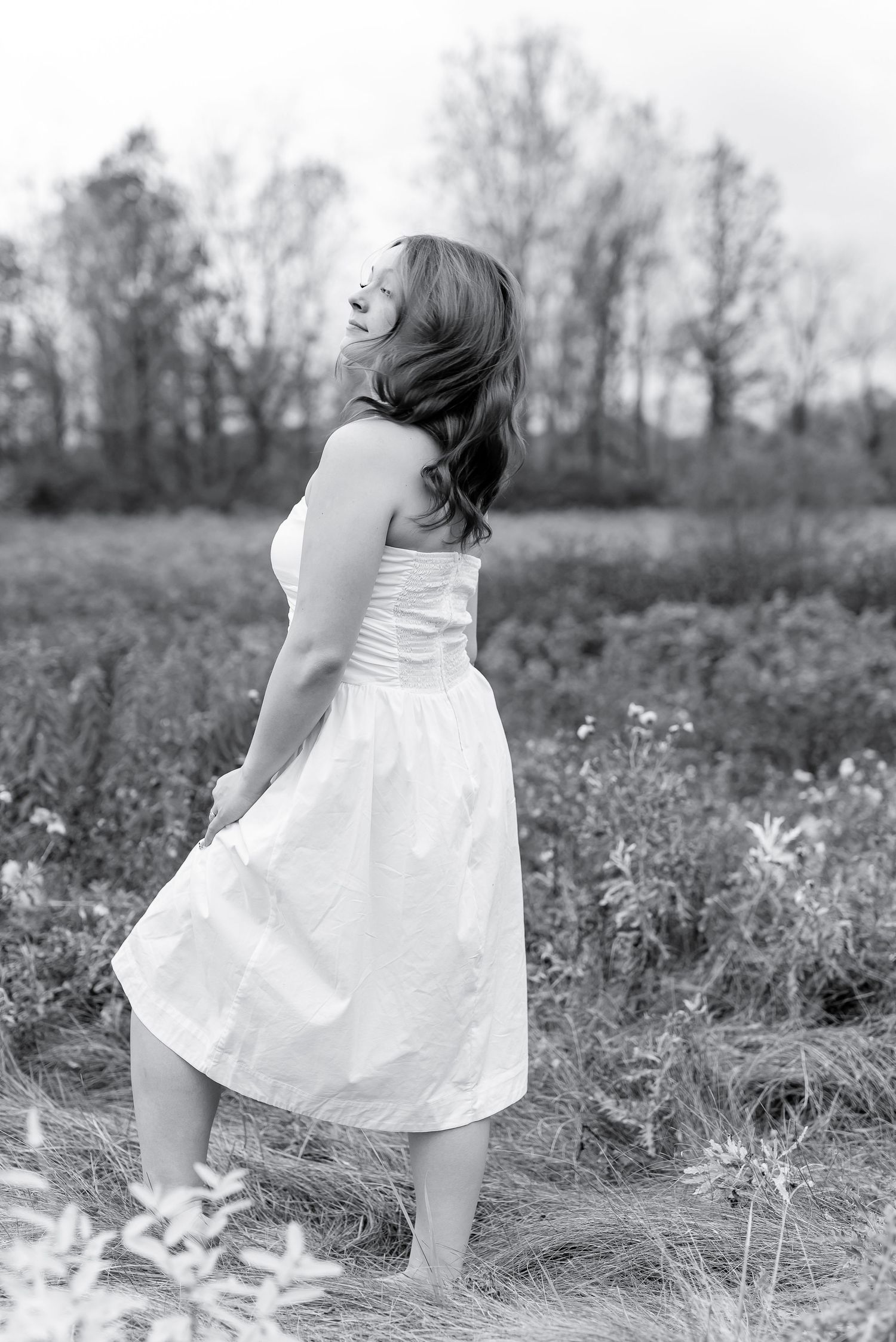Black and white photo of woman in white dress standing in field with trees in background.