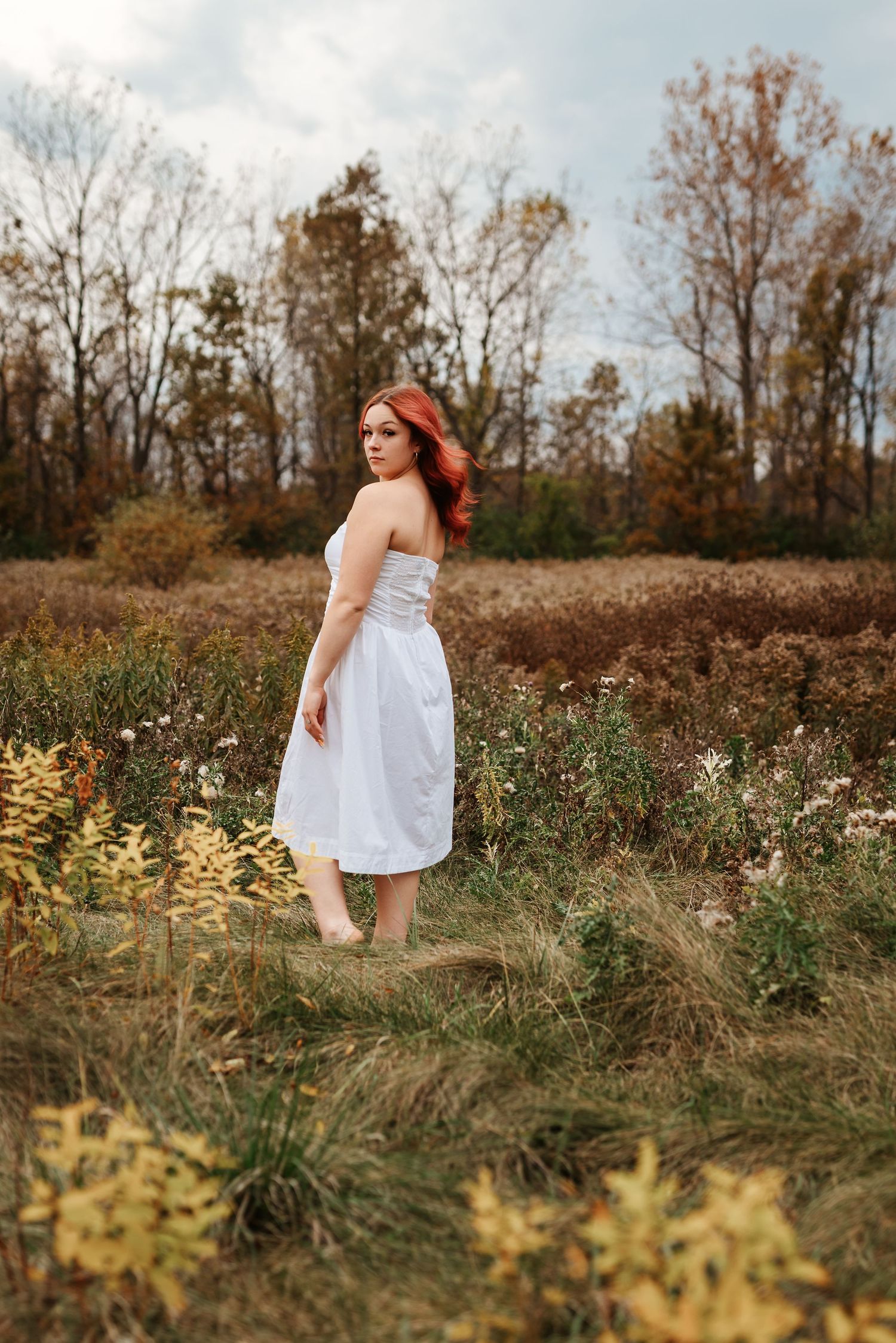 Red-haired woman in white strapless dress standing in autumn field with colorful foliage.