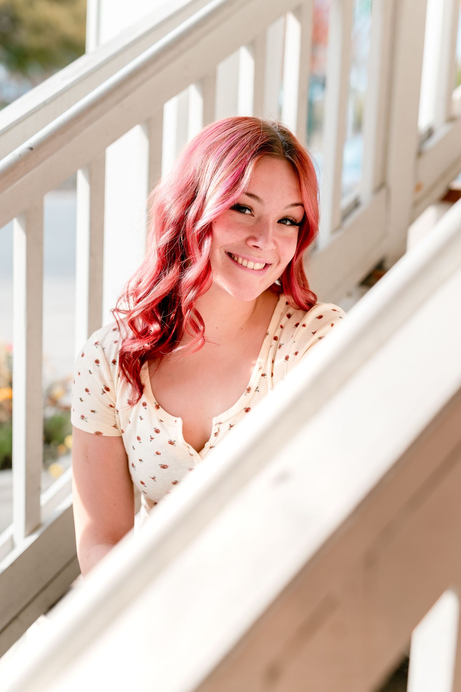 Young woman with pink wavy hair smiles on white staircase railing wearing a floral dress outdoors.