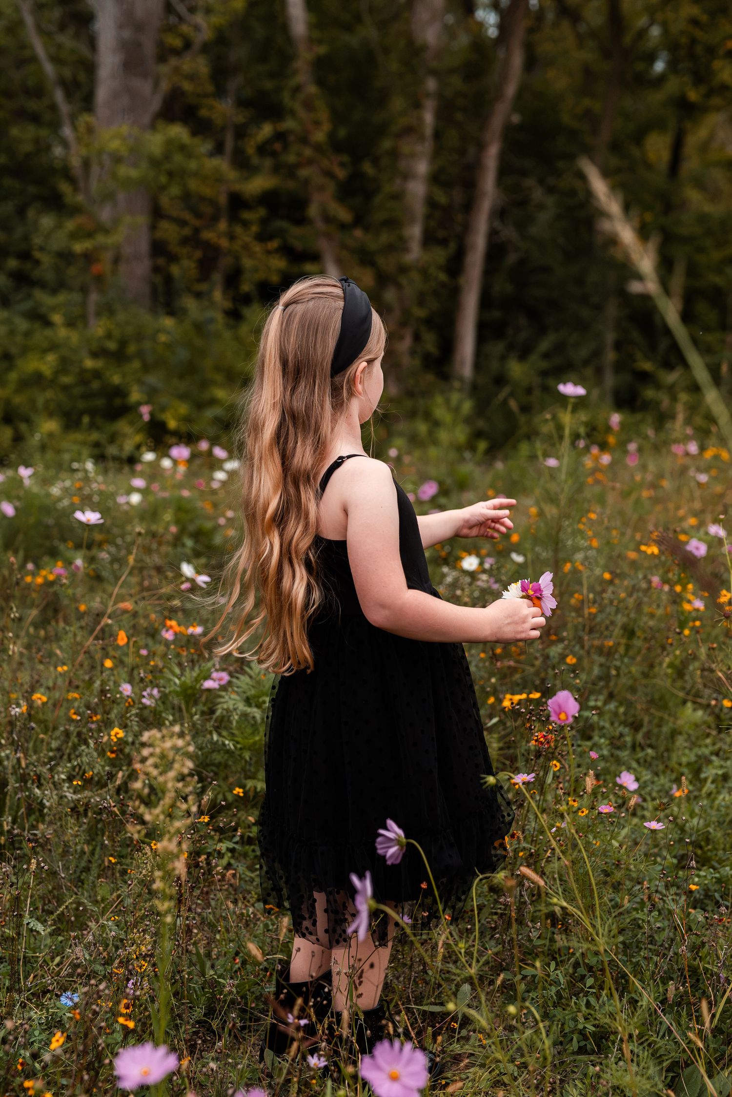Young girl in black dress picking wildflowers in a lush meadow surrounded by colorful blooms and forest trees.