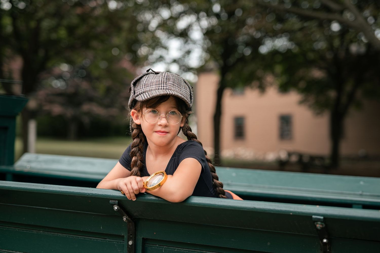 Young girl in plaid cap and braids smiling on a green park bench outdoors.