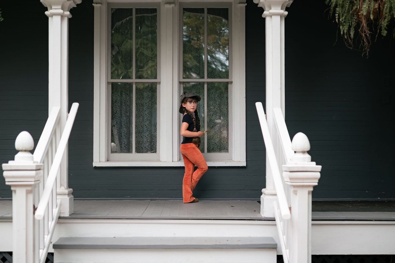 Person in orange pants and black top standing on a white-railed porch of a dark green Victorian house.