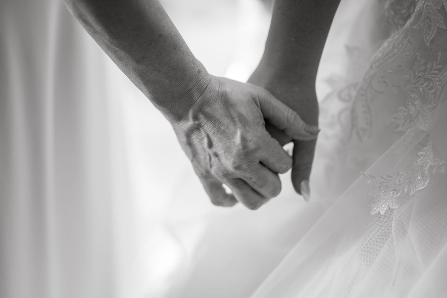 Black and white close-up of newlyweds holding hands against soft window light.