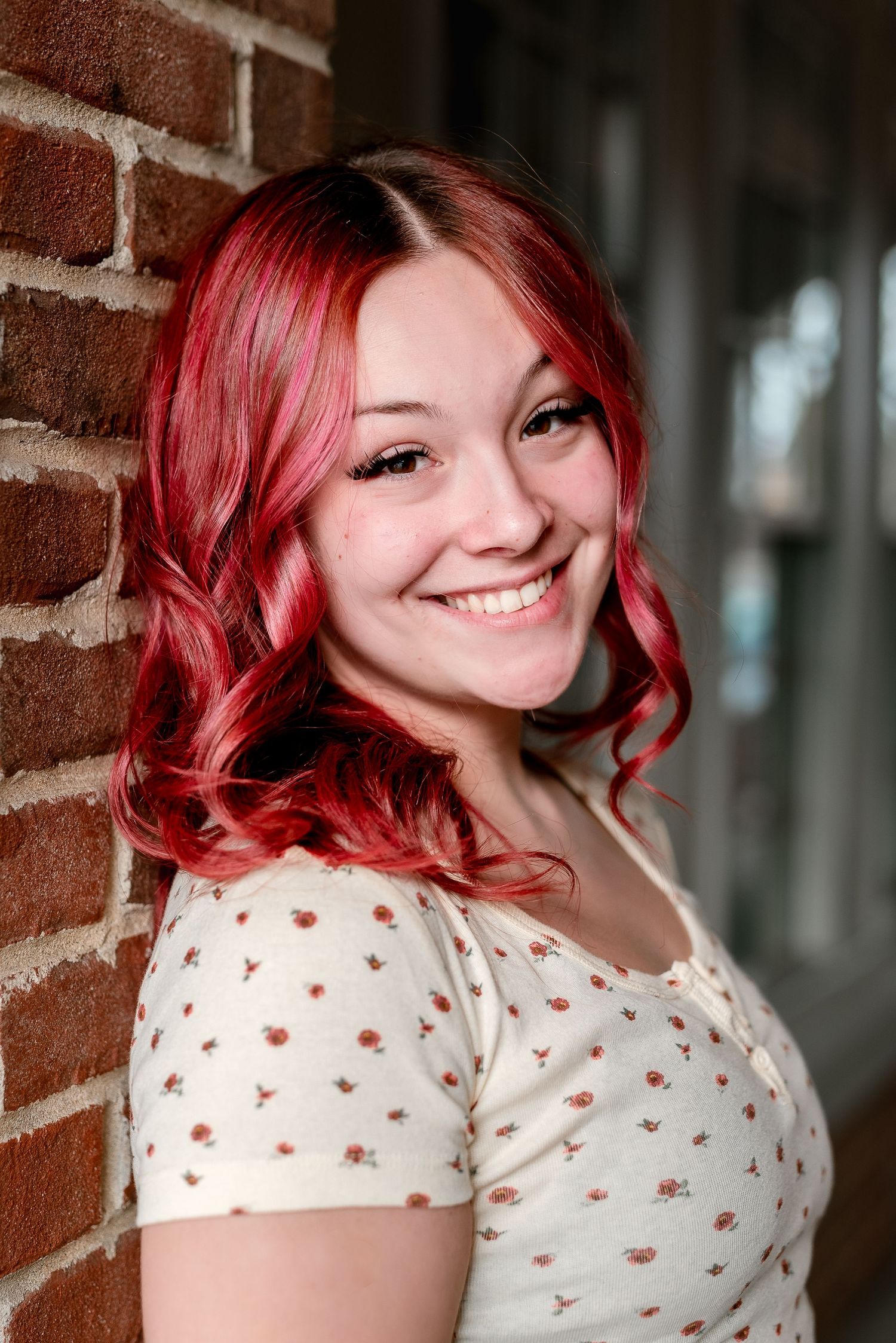 Young woman with vibrant red hair smiling against a brick wall wearing a floral cream top.