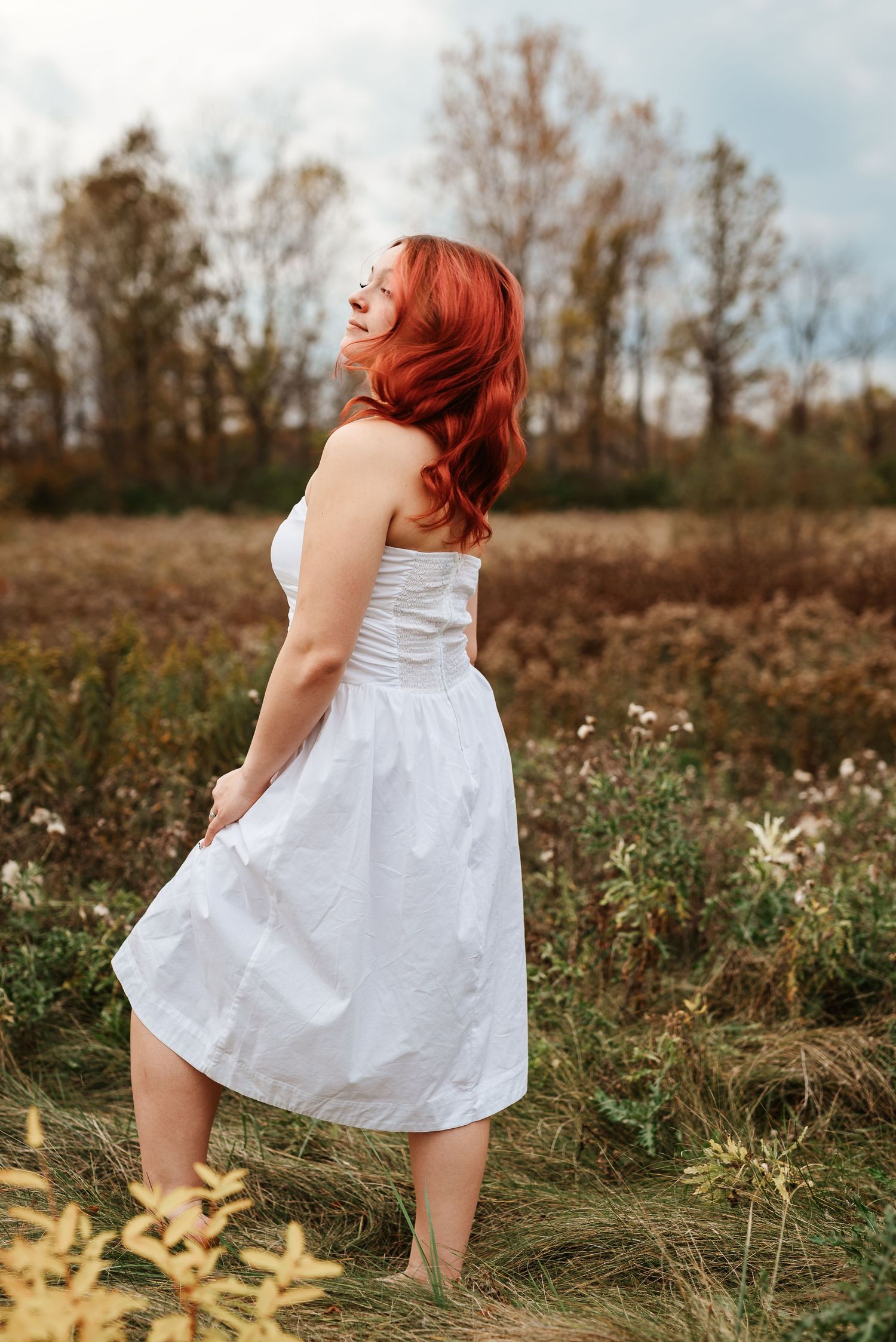 A woman with red hair in a white dress stands in a field, looking up at a cloudy sky.