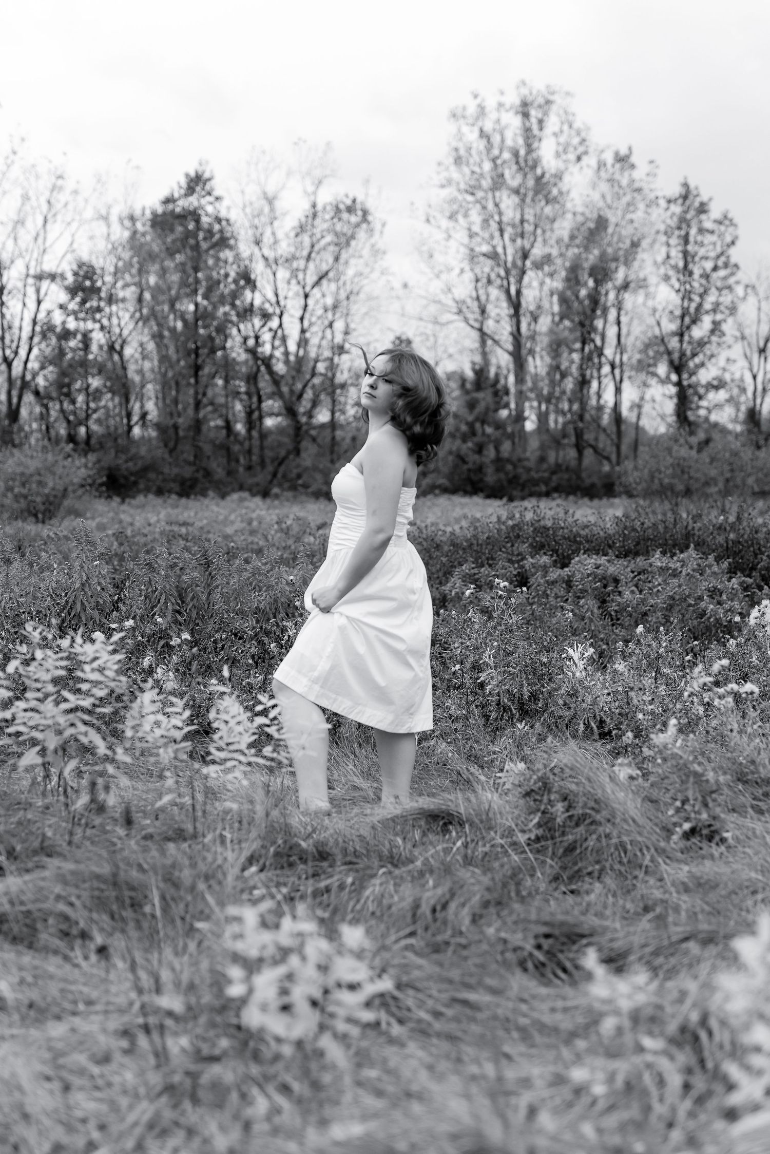 Black and white photo of a woman in a white dress standing in a lush green field surrounded by trees.