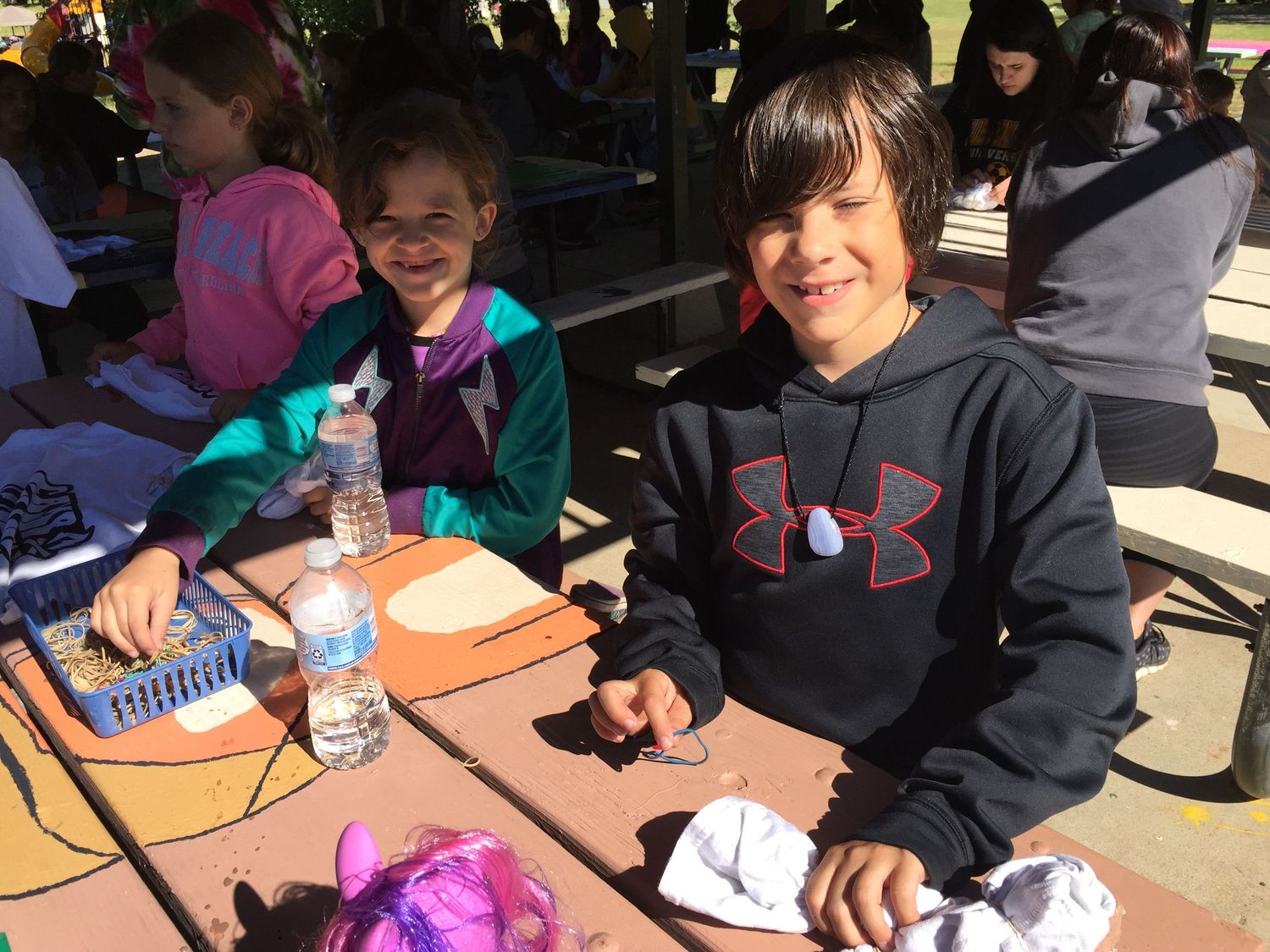 Two children smiling at a picnic table outdoors, one wearing an Under Armour hoodie, enjoying a school lunch break.