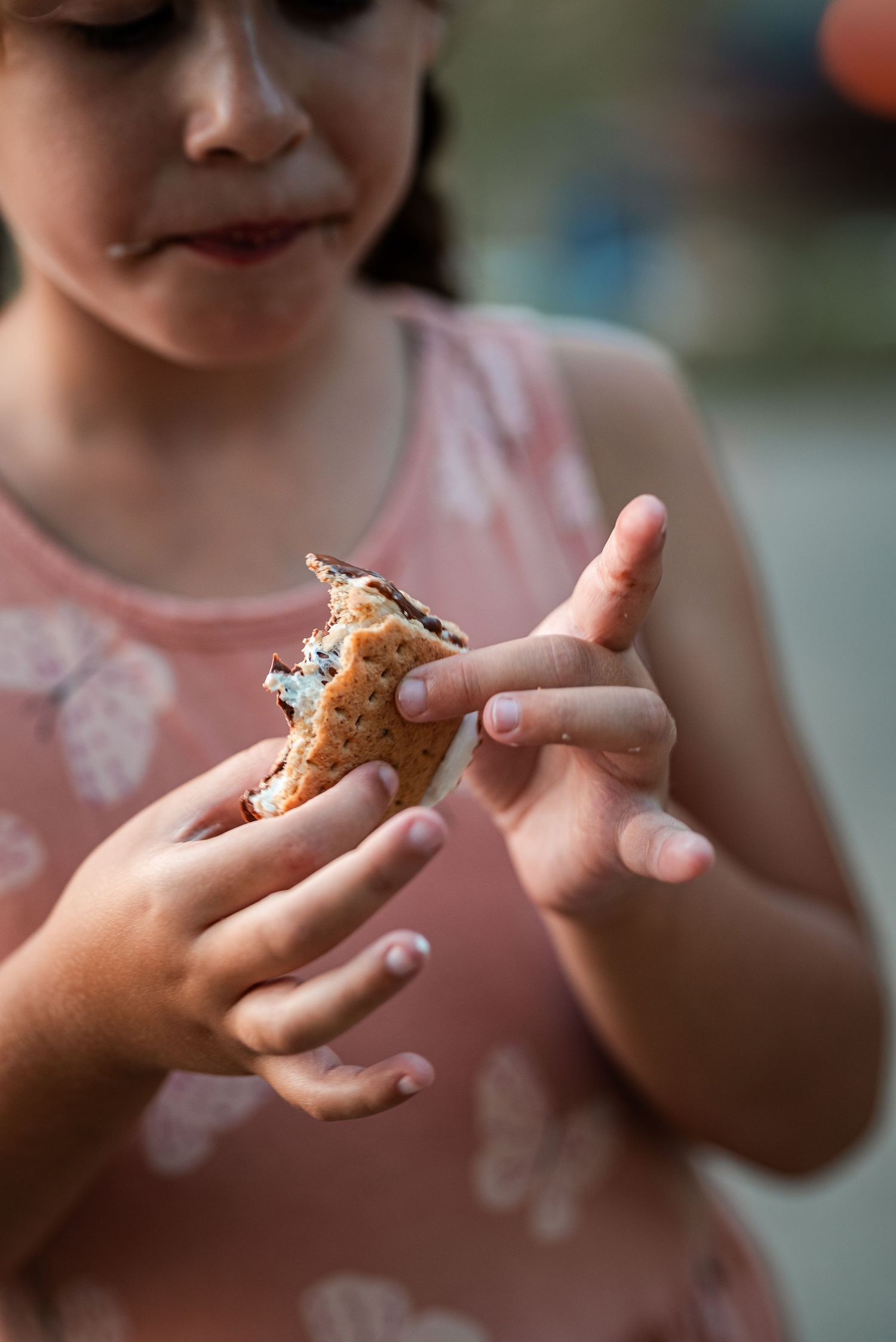 A young girl in a pink dress holds and examines a half-eaten smore with sticky fingers.