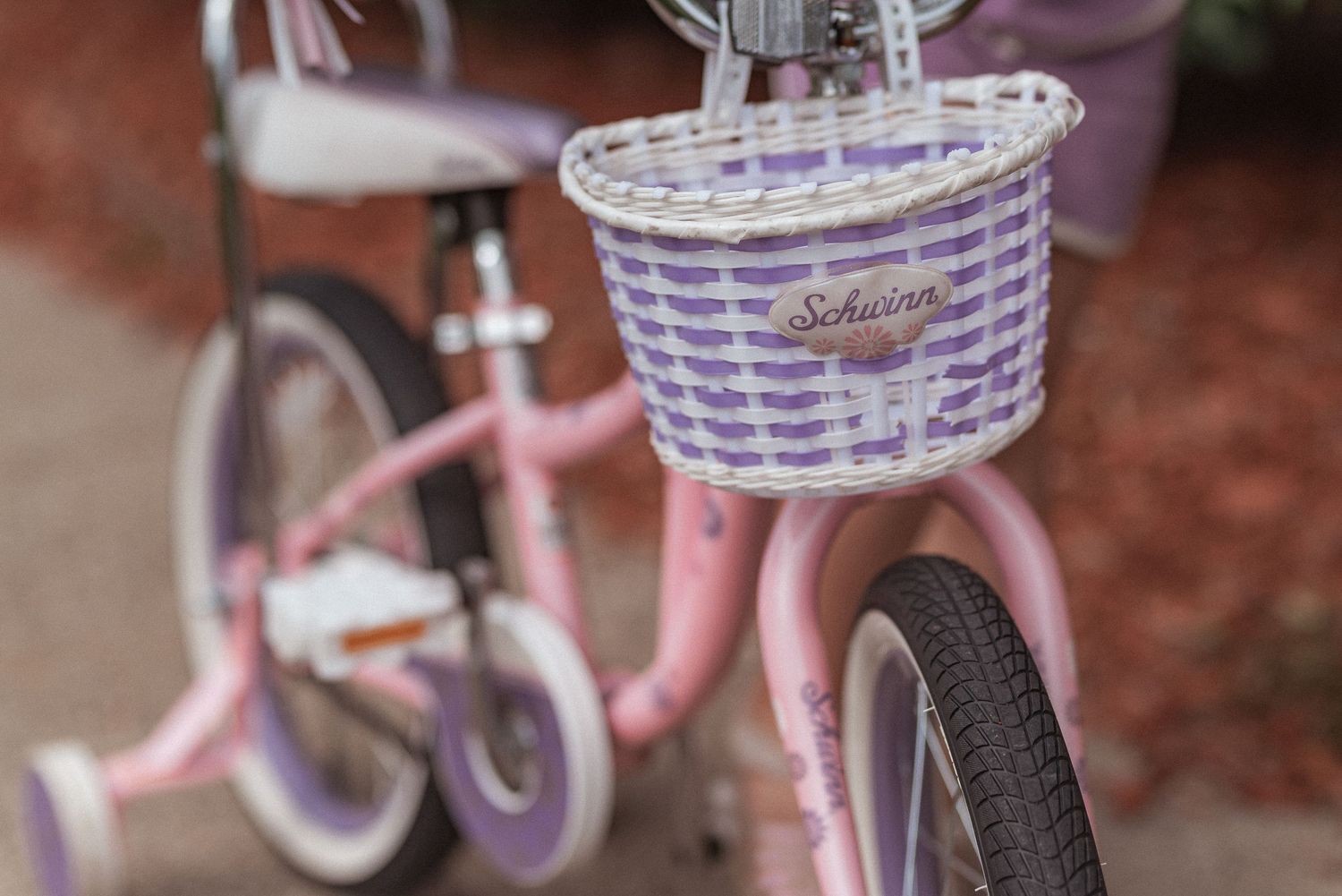 A pink bicycle with a purple striped woven basket mounted on the handlebars, shown in close-up detail.