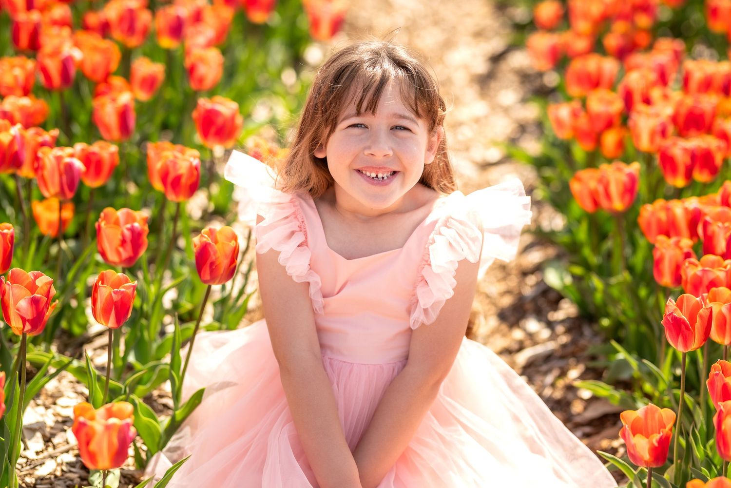 Young girl in pink dress smiling among vibrant red and orange tulips in a sunny flower field.