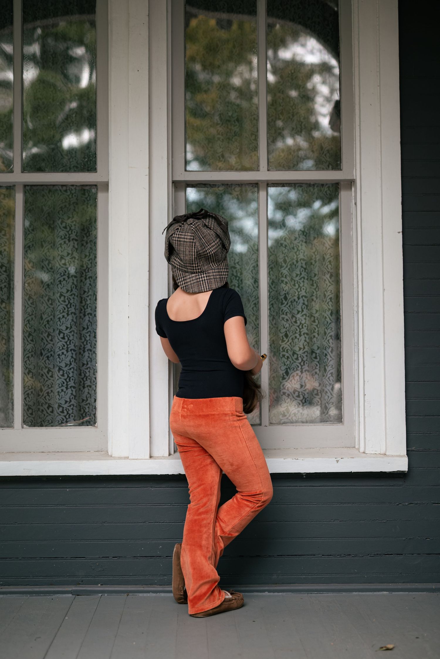 Woman in orange flare pants, black top, and plaid hat stands with back turned, peering through a large window.